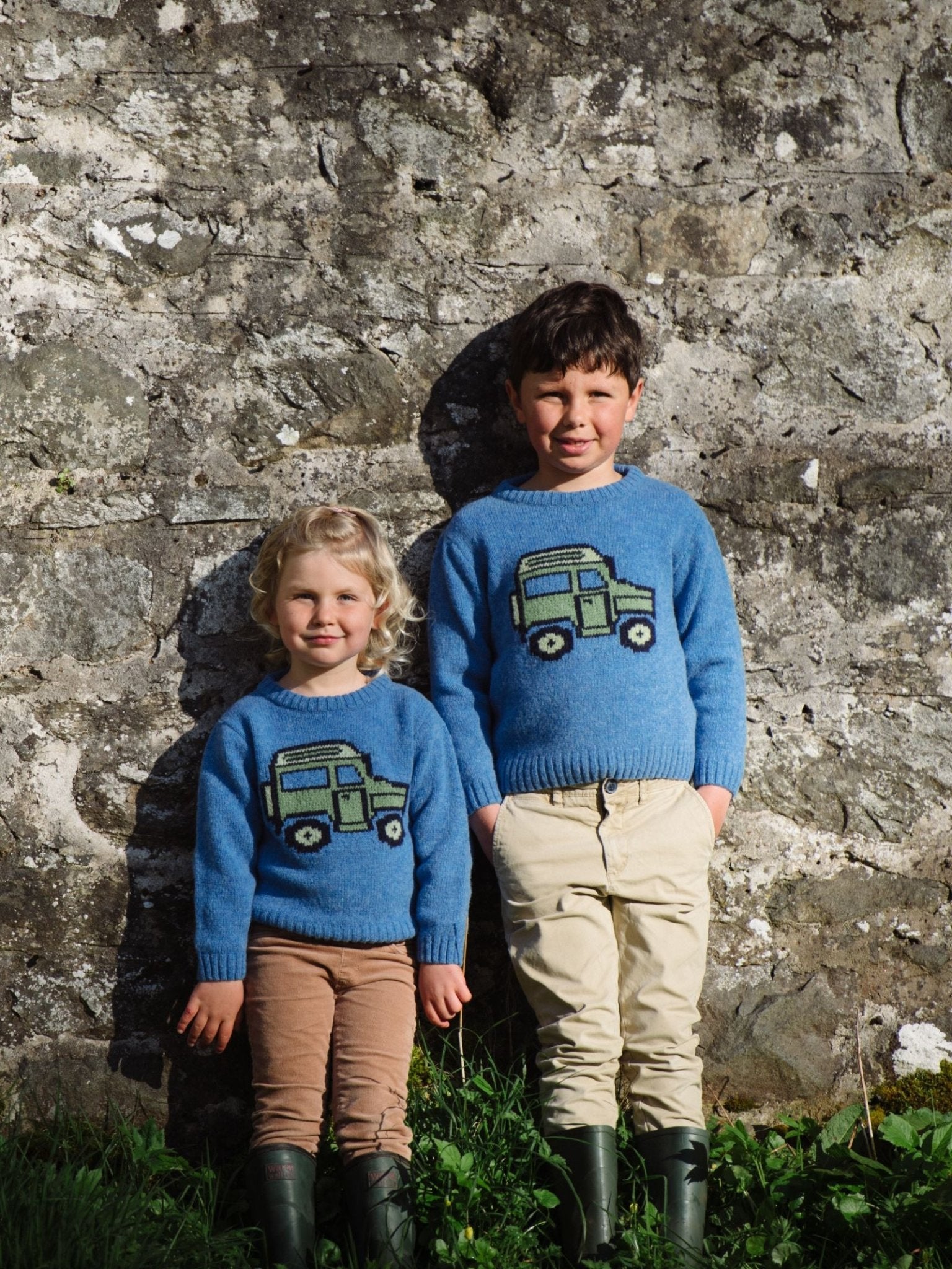 Two young children smile in front of a stone wall, both wearing Campbells of Beauly Mini Land Rover Defender Jumpers featuring a green truck design. The older child has hands in pockets; the younger stands with arms by their sides.