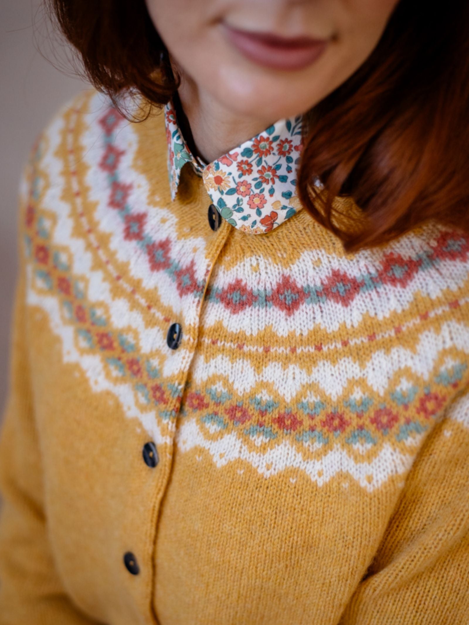 A red-haired woman wears the Campbells of Beauly Shetland Fairisle Cardigan, a mustard yellow Pure New Wool knit with black buttons, layered over a colorful floral collared shirt. Her relaxed face is partially visible.