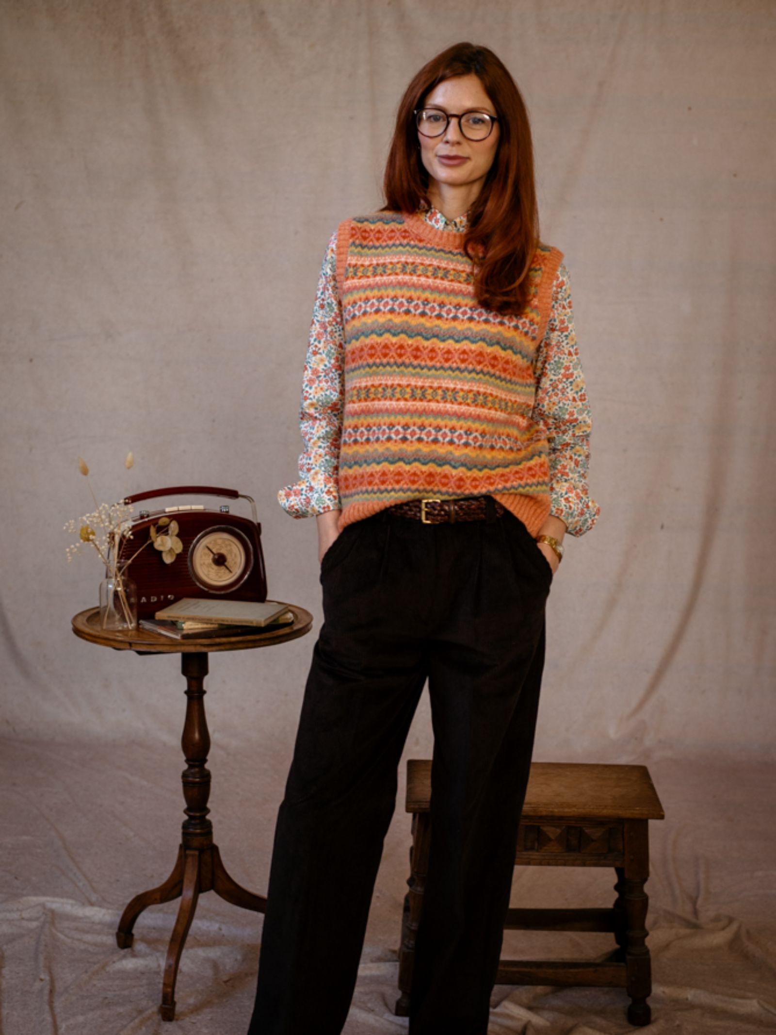 A woman with long brown hair and glasses stands indoors, wearing a Campbells of Beauly Fairisle Crew Tank over a floral shirt and black pants. Behind her are a small round table with a vintage radio, dried flowers, and a wooden stool.