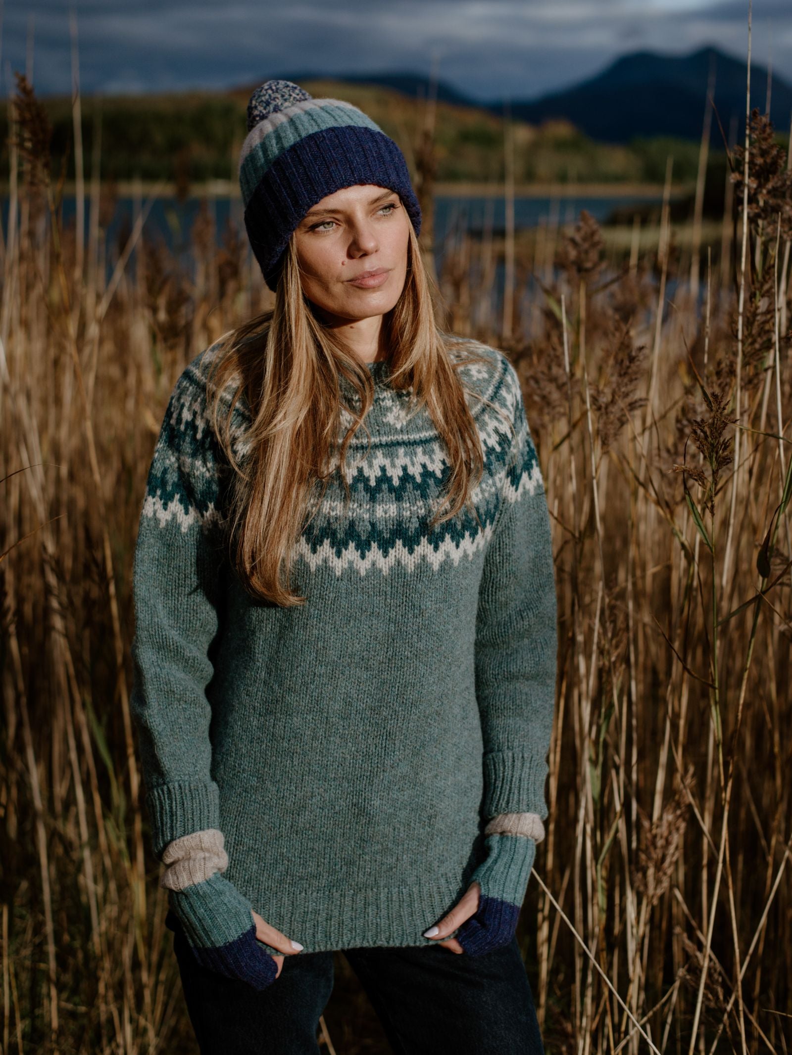 A woman stands outdoors in tall grass, with Scottish Borders mountains and lakes behind her, wearing Campbells of Beauly Stripe Wristwarmers, a matching beanie, and a knitted blue and green lambswool sweater under a cloudy sky.