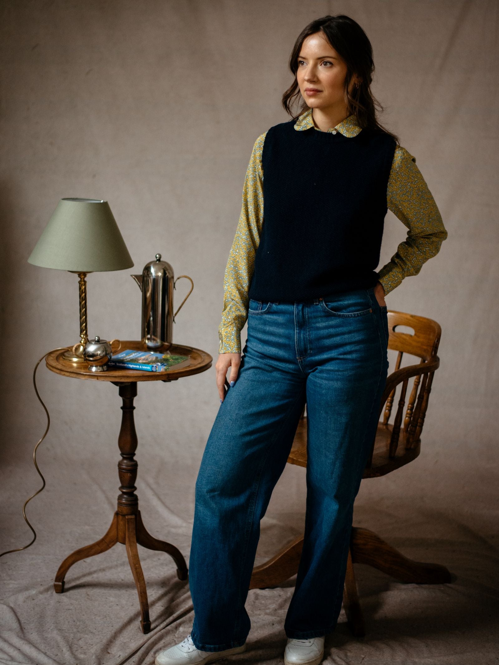 A woman in a patterned blouse, Campbells of Beauly Moss Stitch Crew Tank, and blue jeans stands by a round wooden table with a lamp, small tray, and French press; a wooden chair is behind her against a neutral backdrop.