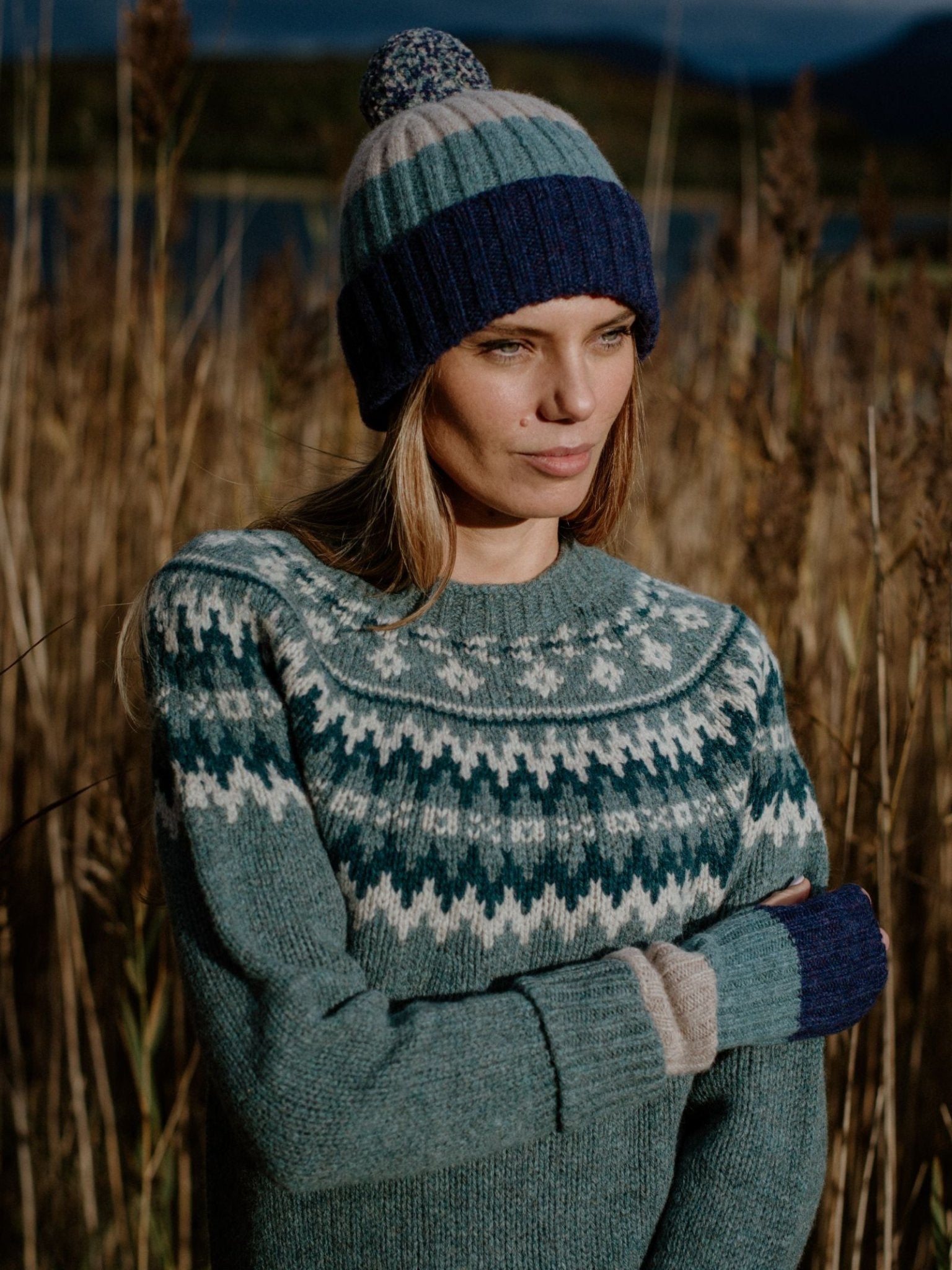 A woman stands in a field, wearing the Campbells of Beauly Lambswool Stripe Hat with Pom-Pom. She gazes thoughtfully to the side, capturing the tranquil charm of the Scottish Borders.