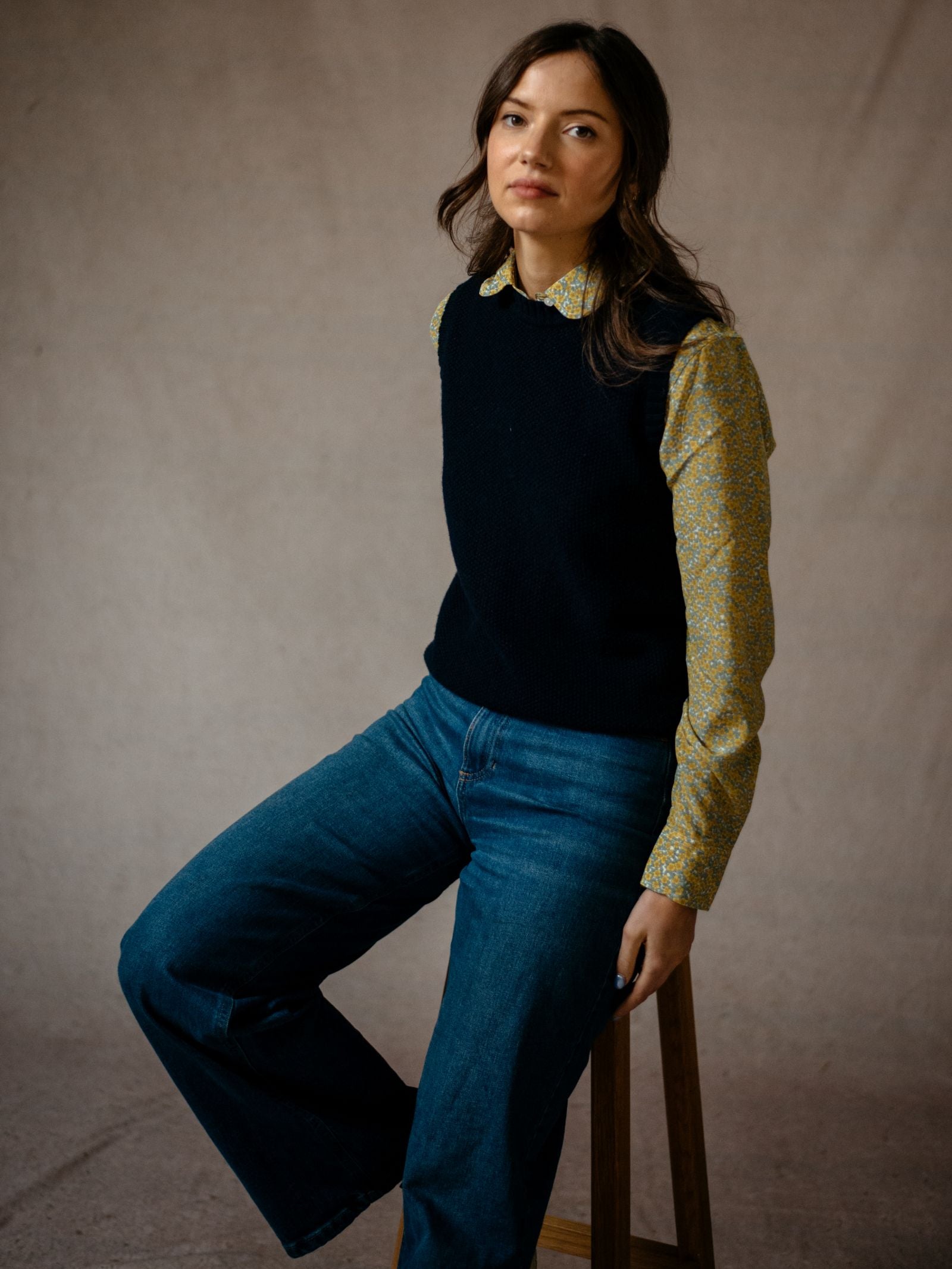 A woman with long dark hair sits on a wooden stool, wearing blue jeans and a yellow patterned shirt layered under the Campbells of Beauly Moss Stitch Crew Tank. She faces the camera against a beige background, highlighting this versatile piece.