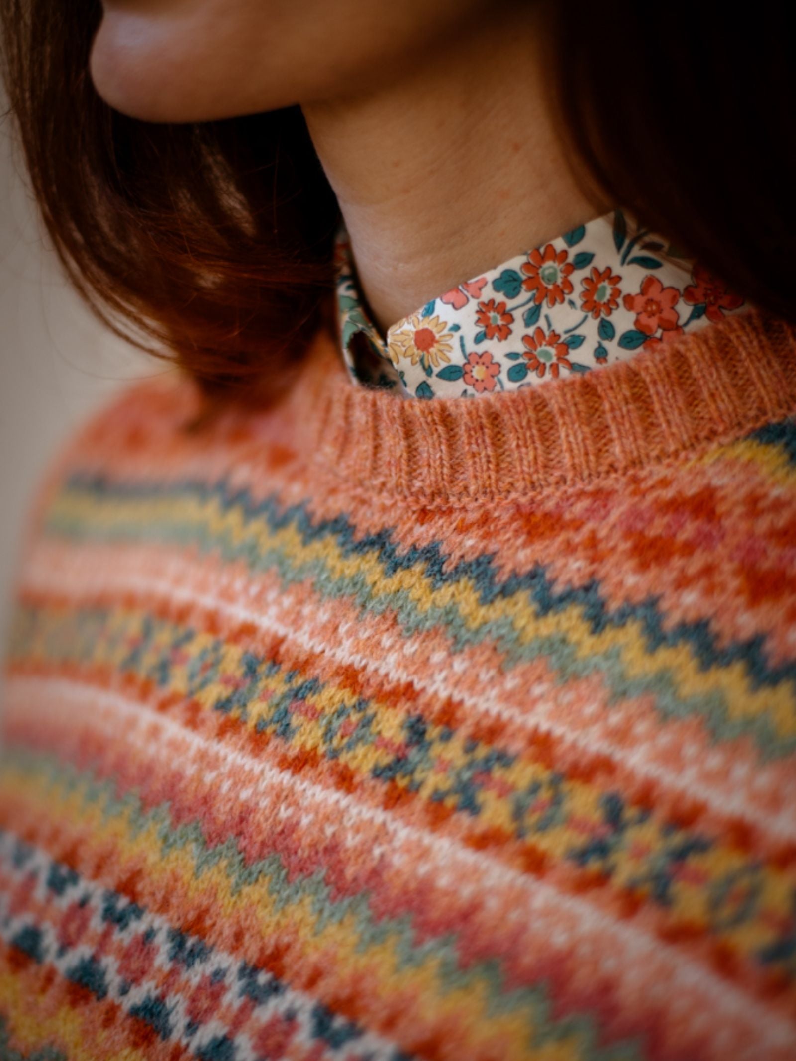 A close-up of a person with shoulder-length brown hair wearing the Campbells of Beauly Fairisle Crew Tank, featuring a traditional pattern over a floral-collared shirt. Only the lower face, neck, and upper torso are visible.