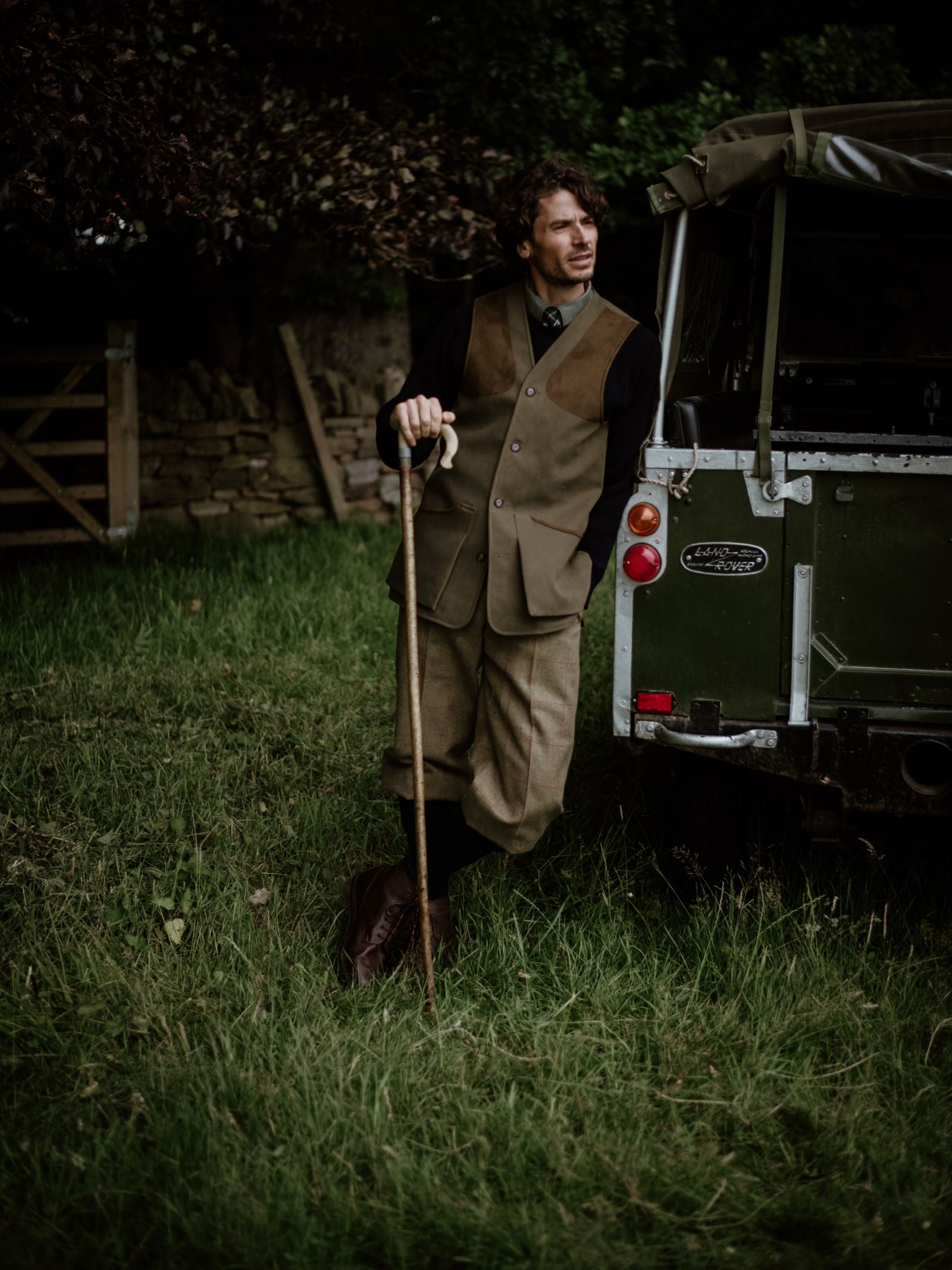 A man in country attire and a Campbells of Beauly Cotton Shooting Waistcoat leans on a walking stick beside a green Land Rover on grass, with trees and a wooden fence in the background.