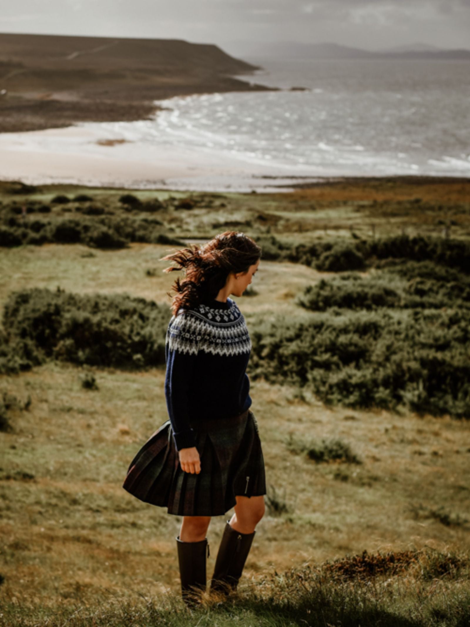 A woman, her hair blowing in the wind, stands on a grassy hillside by a cloudy coast wearing the Campbell's of Beauly Tweed Kilt with boots and a patterned sweater, gazing toward waves and distant hills.