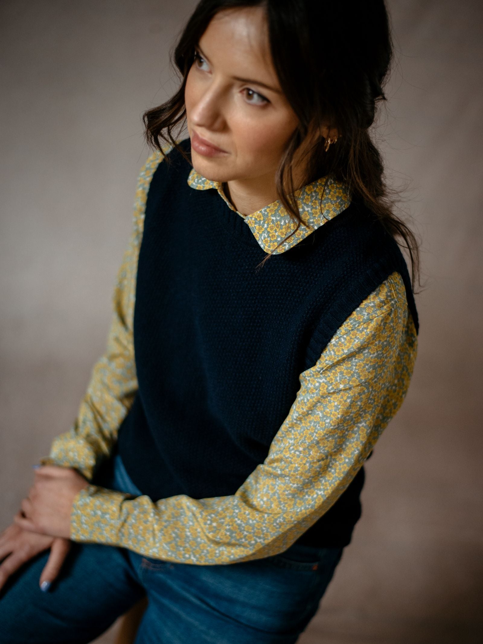 A woman with wavy brown hair sits sideways, hands on her lap, wearing a yellow floral shirt under the Campbells of Beauly Moss Stitch Crew Tank and blue jeans.