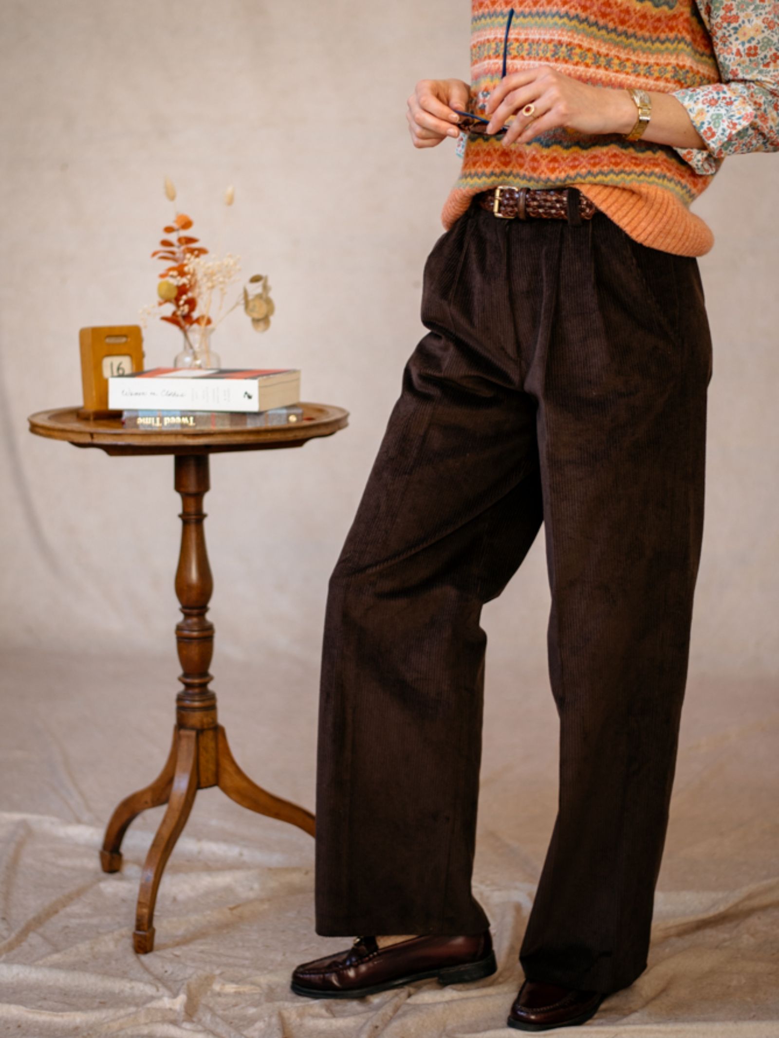 A person in a patterned sweater vest, floral shirt, and Campbells of Beauly Wide Leg Cords stands beside a small round wooden table with books, a decorative vase, and dried flowers. Their face is not shown.