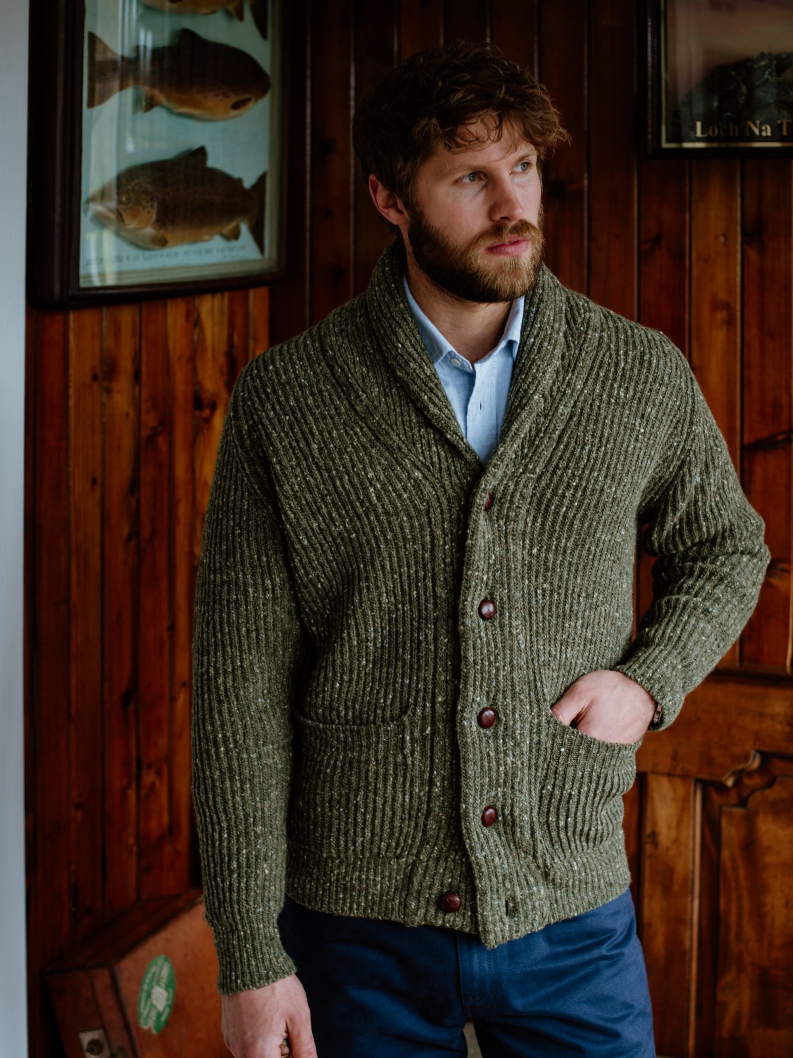 A bearded man stands indoors, wearing a Campbells of Beauly Donegal Lambswool Shawl Collar Cardigan with leather buttons over a light blue shirt, one hand in his pocket. Wooden walls, framed fish art, and a door are in the background.