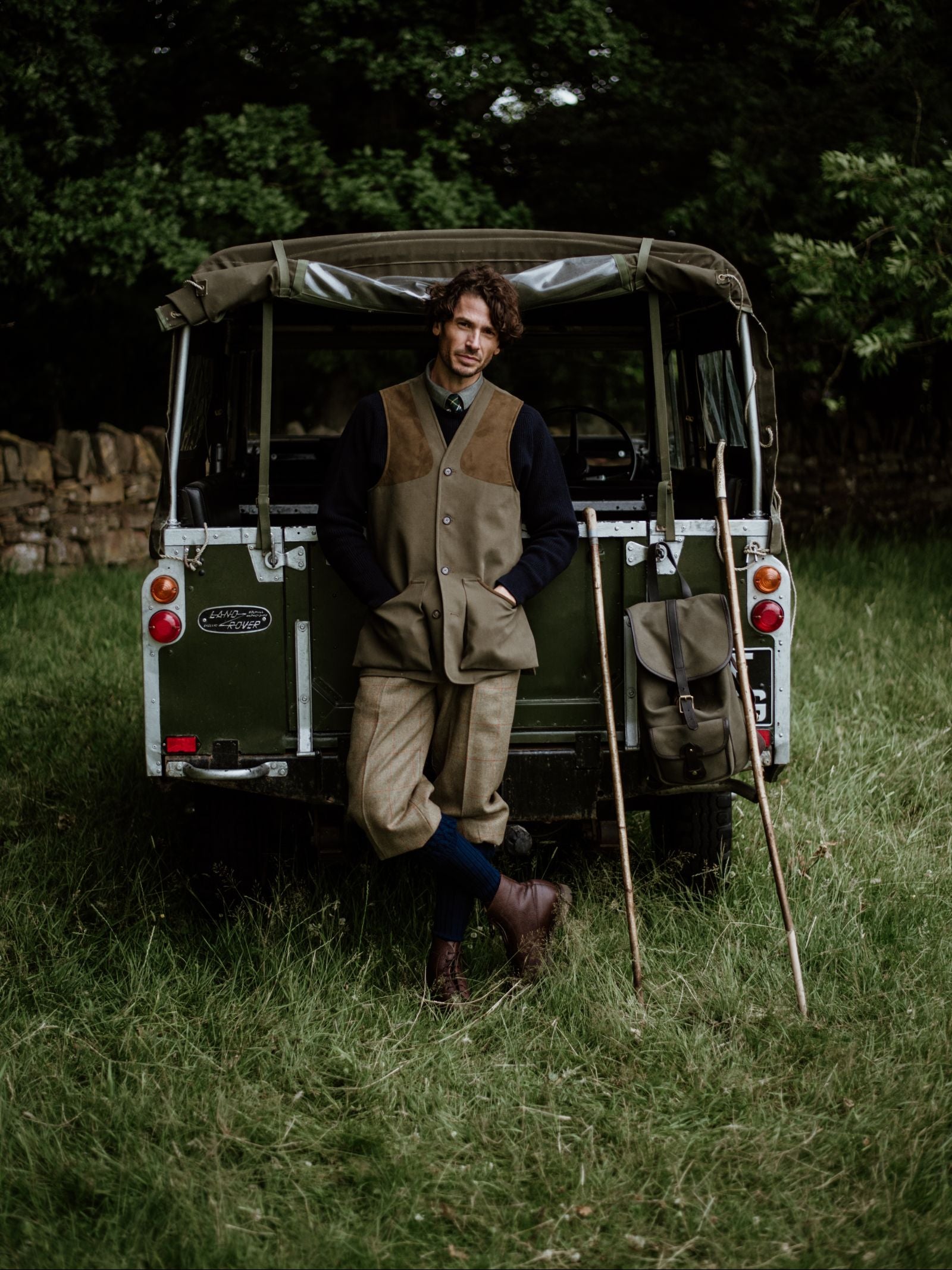A man in vintage outdoor clothing leans against a green Land Rover on grass, his Campbells of Beauly Cotton Shooting Waistcoat, two wooden walking sticks, and satchel evoking classic countryside pursuits among the trees.