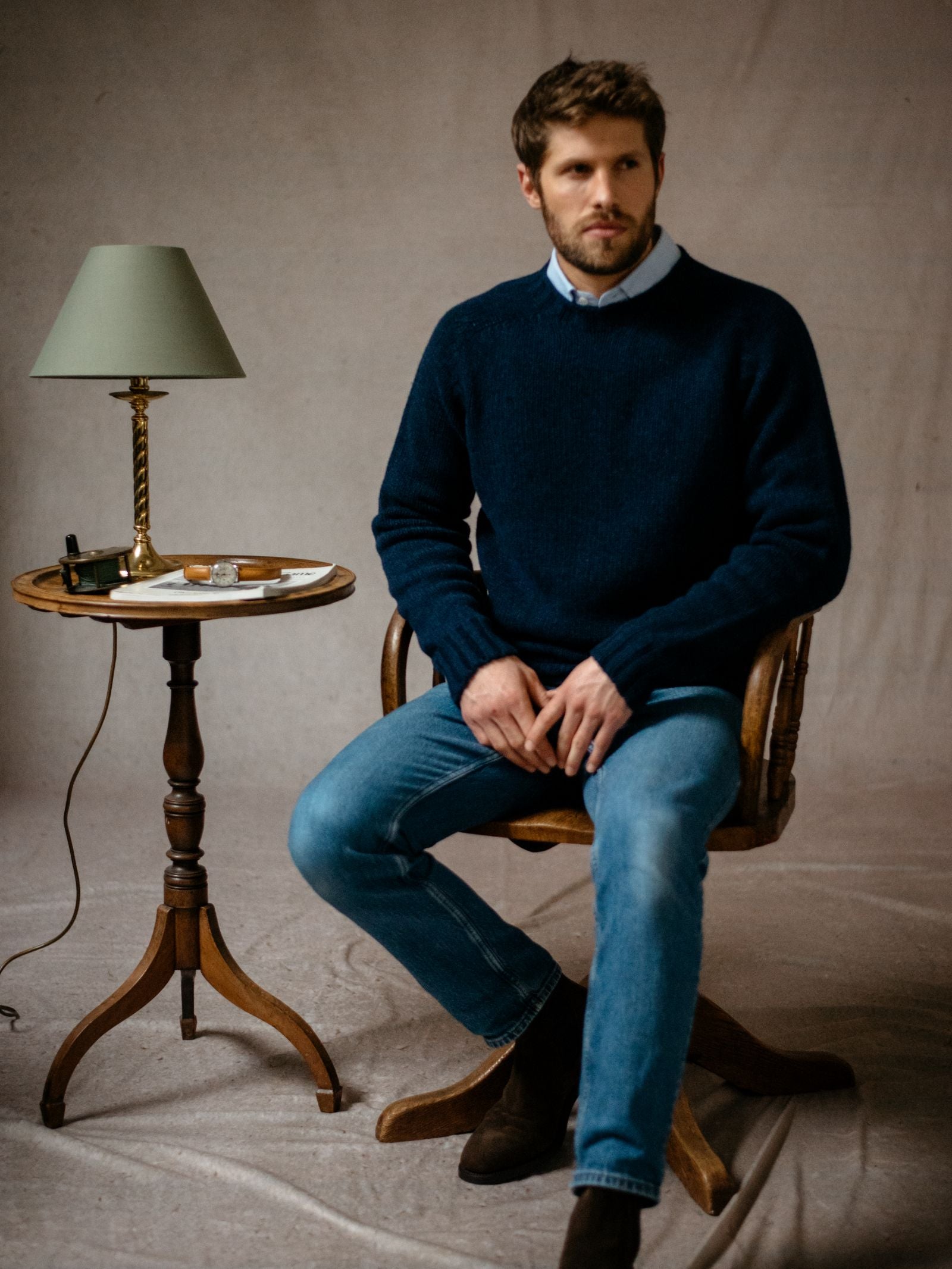 A man with short brown hair and a beard sits on a wooden chair, wearing a navy Geelong Lambswool Chunky Crew Jumper by Campbells of Beauly over a blue shirt and jeans. Beside him is a round table with a green lamp, pipe, matchbox, and white dish.