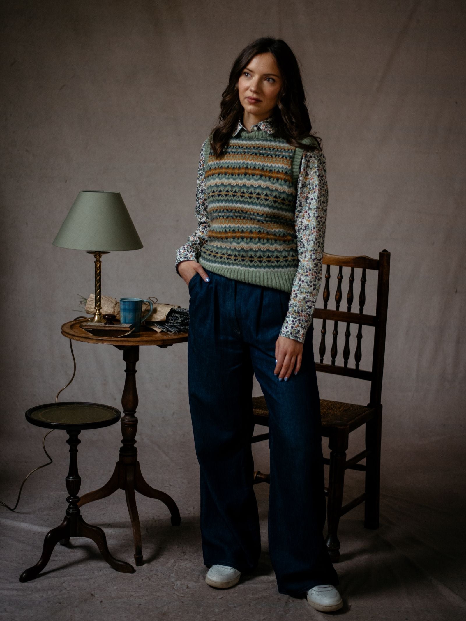 A woman in wide-leg jeans, a floral blouse, and the Campbell's of Beauly Fairisle Crew Tank stands by a wooden table with a lamp and teacup in a softly lit room with neutral tones.