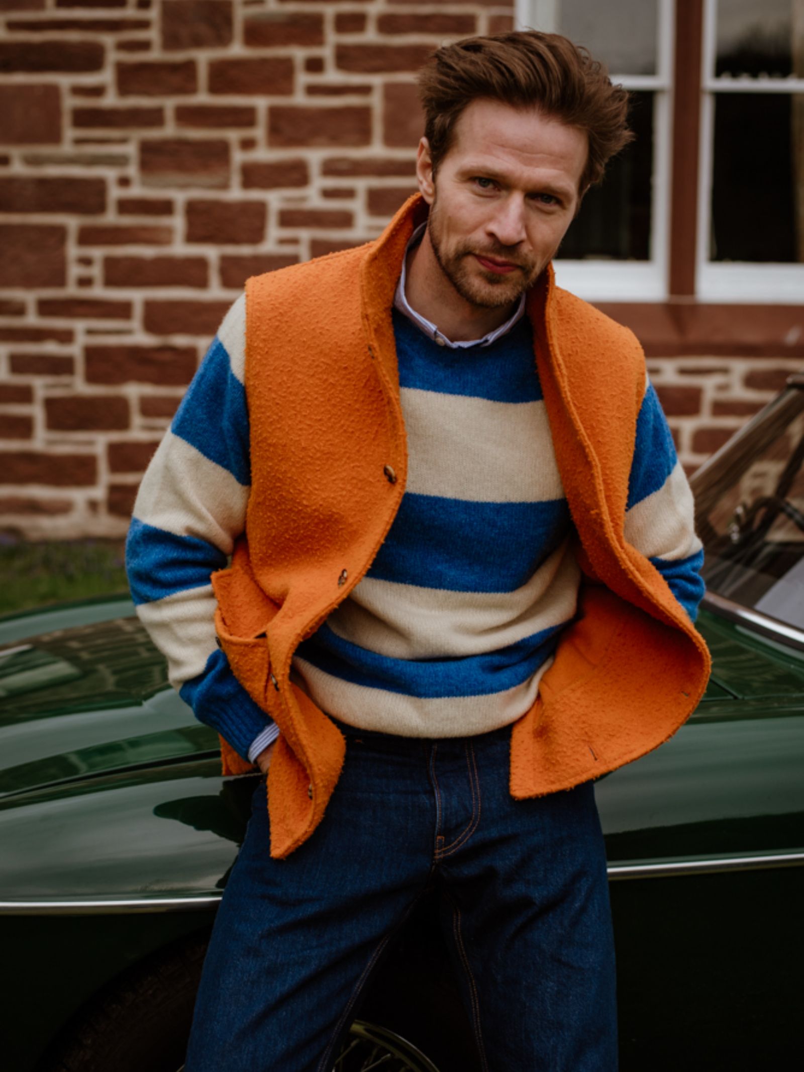 A man with brown hair, in a blue and white striped sweater, jeans, and a Campbells of Beauly Casetino Fleece Gilet, leans on a dark green car. A brick building with white-trimmed windows appears in the background.