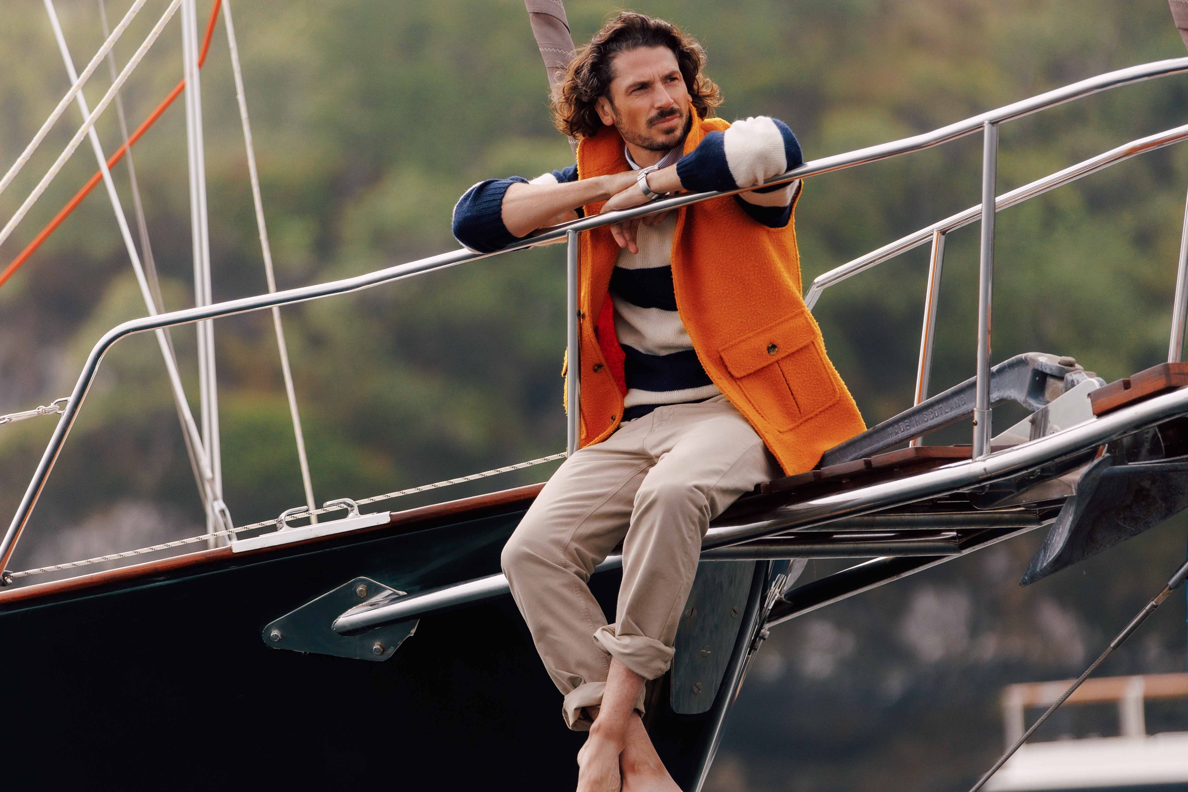 A man with curly hair wearing a striped sweater, orange vest, and beige pants sits barefoot on the edge of a sailboat, resting his arms on the railing, with a blurred green background.