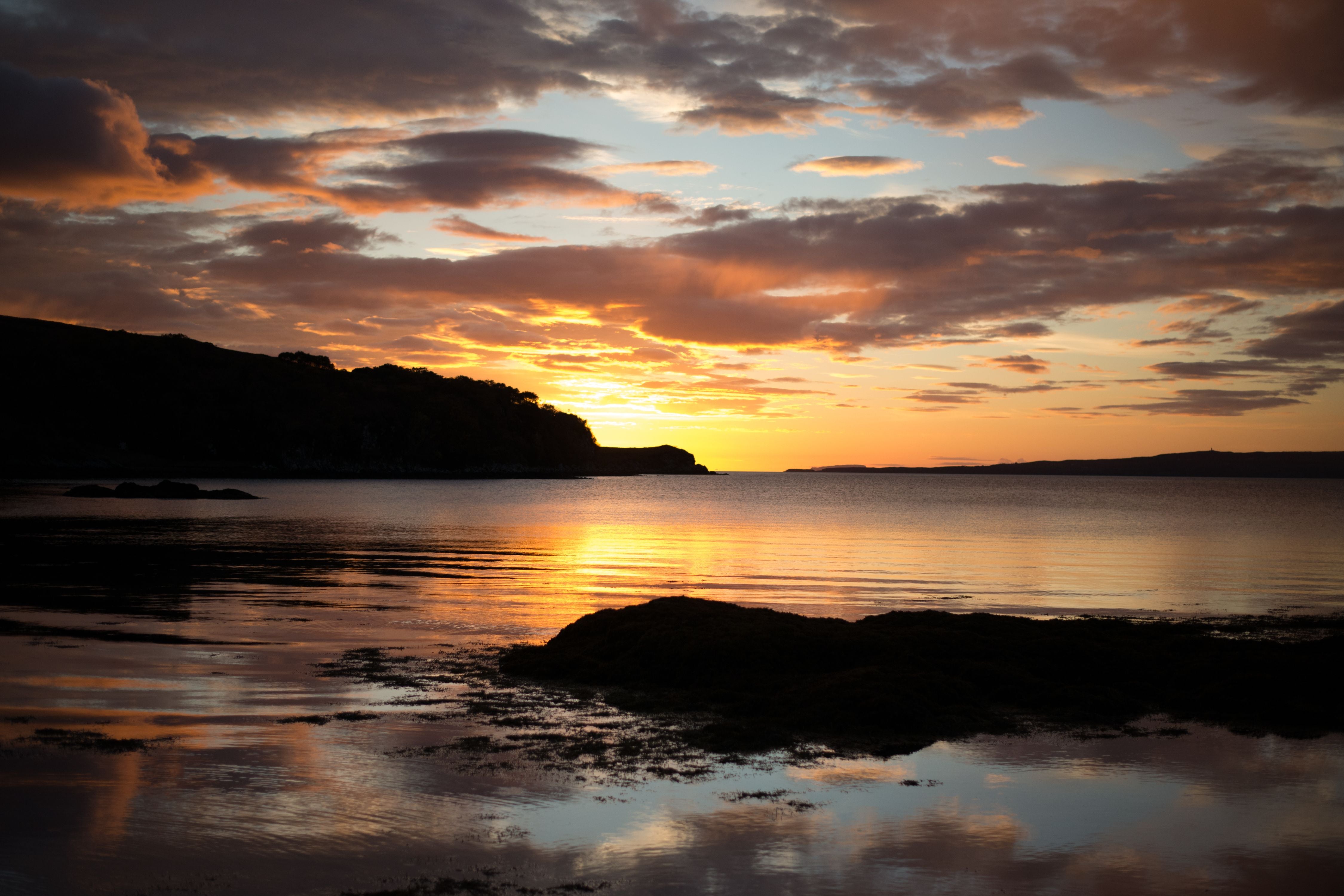 A tranquil sunset over a calm lake, with dark silhouettes of hills and rocks in the foreground. The sky glows with orange, yellow, and purple hues, reflecting beautifully on the water’s surface.