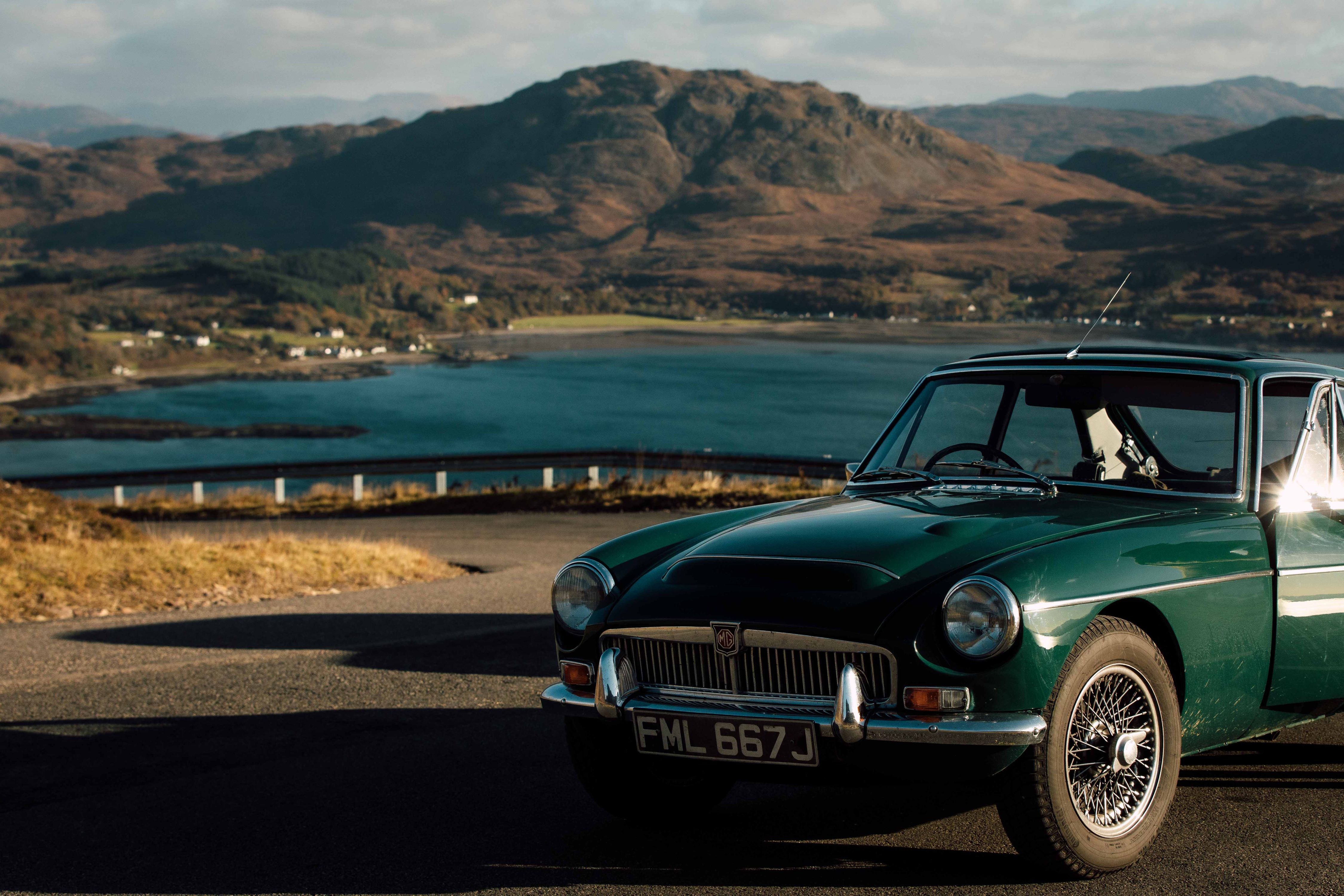 A classic green MG car parked on a scenic road overlooking a blue lake and rugged mountains under a partly cloudy sky.