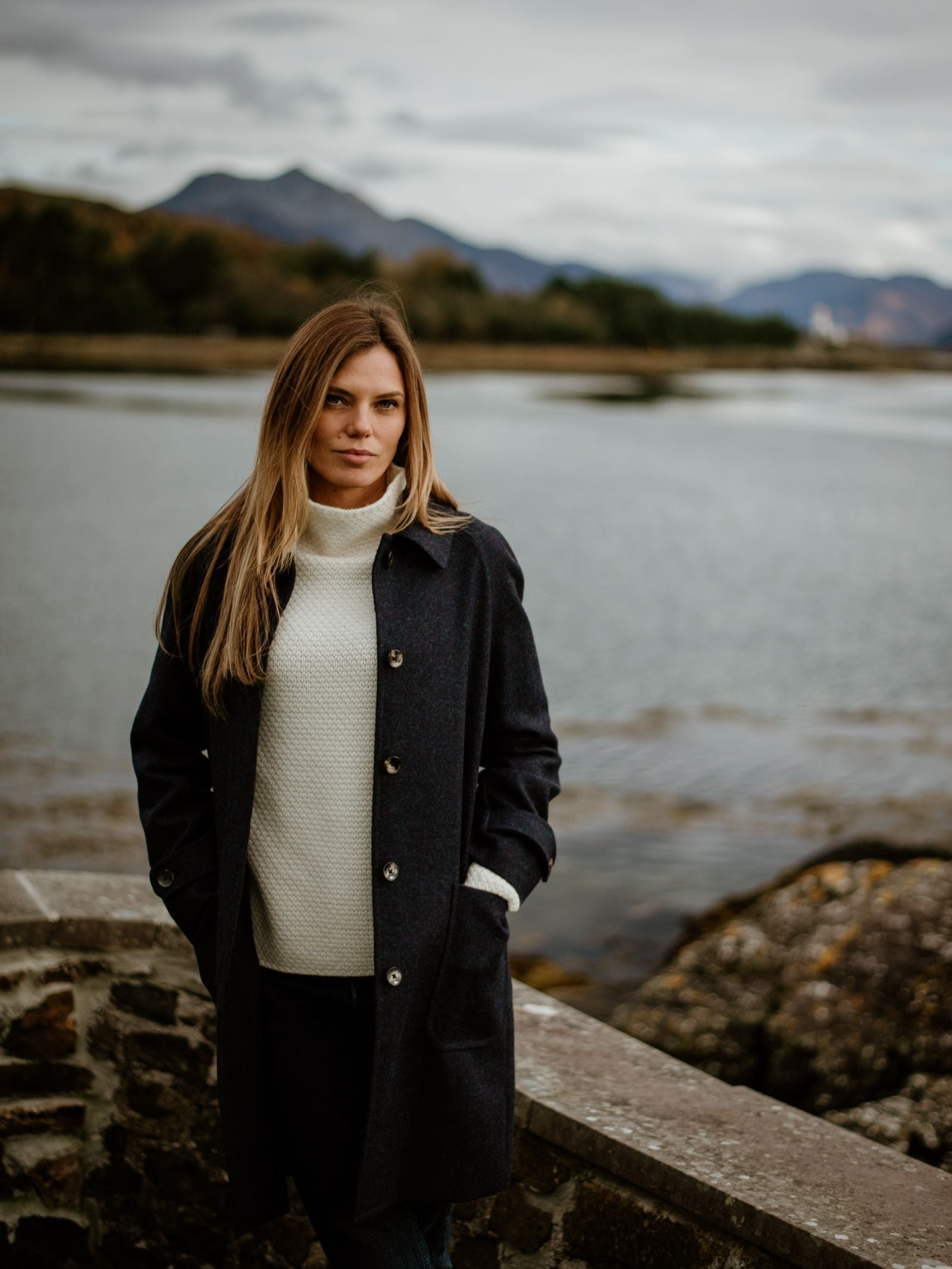 A woman with long hair wears the Campbells of Beauly Midnight Collar Coat and a white sweater, standing by a stone wall near a calm lake, mountains, and cloudy skies.