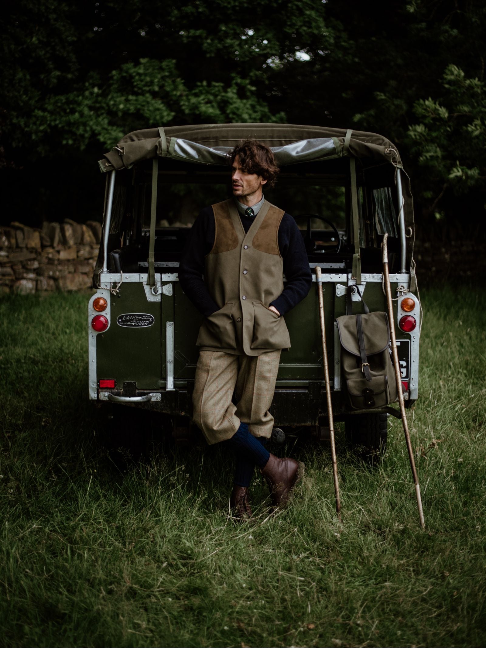 A person in country attire, wearing a Campbells of Beauly Cotton Shooting Waistcoat, leans against an old green Land Rover on grass, with two walking sticks and a bag nearby and trees in the background.