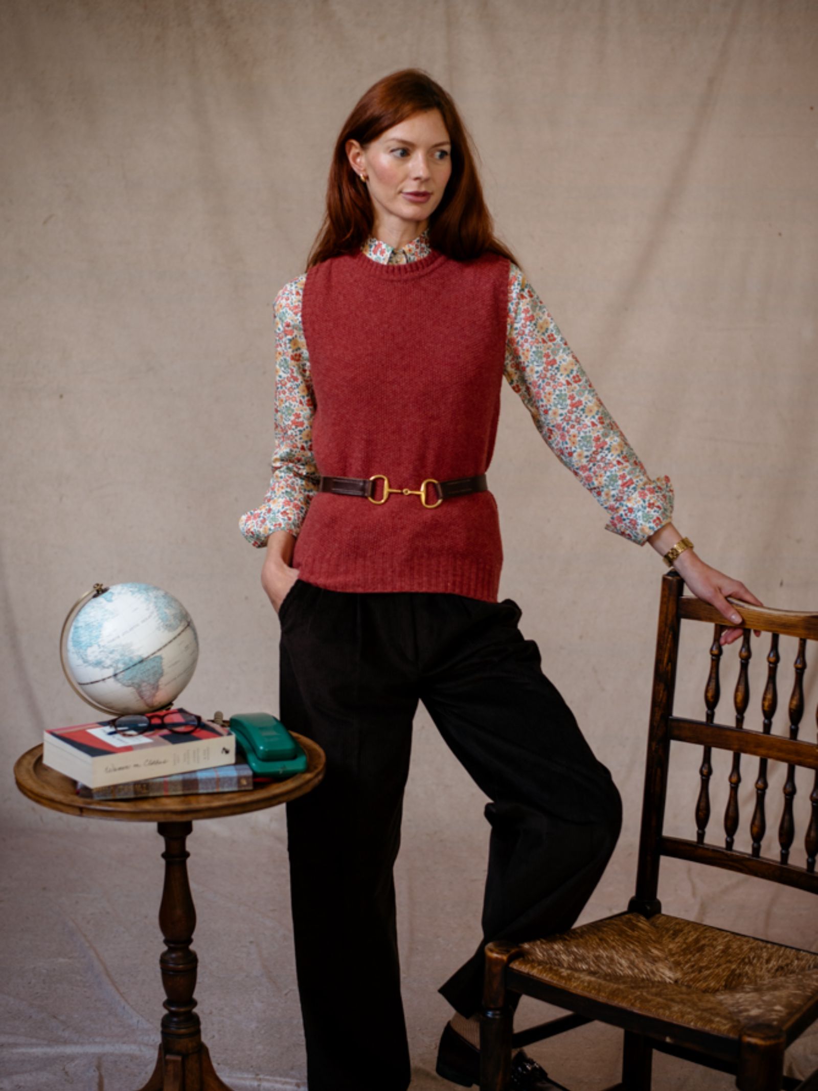 A red-haired woman wears a floral blouse layered with the Moss Stitch Crew Tank by Campbells of Beauly. She stands beside a wooden chair and a small table displaying a globe, books, and a green rotary phone.