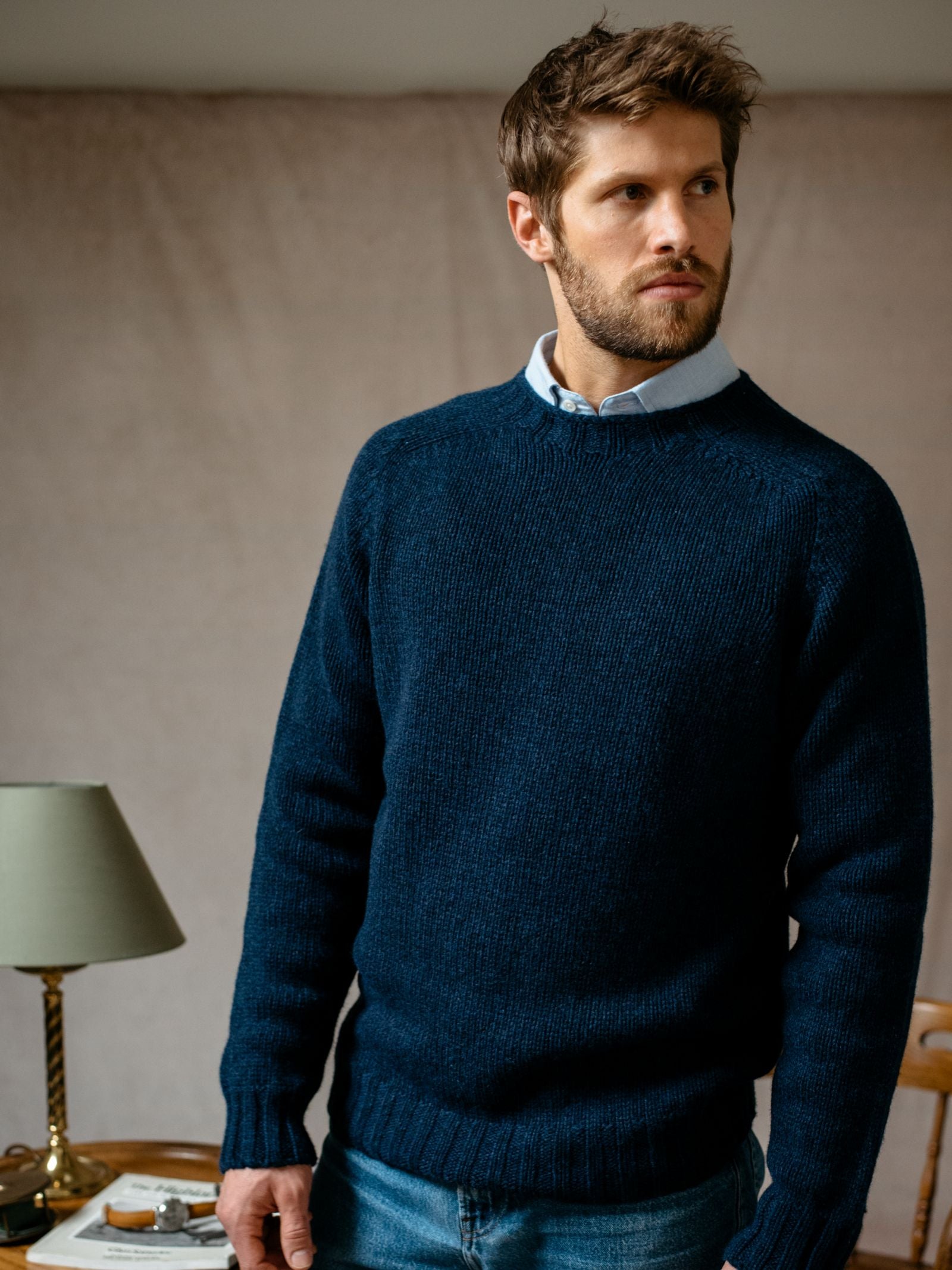 A man with short brown hair and a beard wears a Campbells of Beauly Geelong Lambswool Chunky Crew Jumper in dark blue over a light blue collared shirt and blue jeans. Indoors, he looks left; behind him are a beige wall, green lamp, and table.