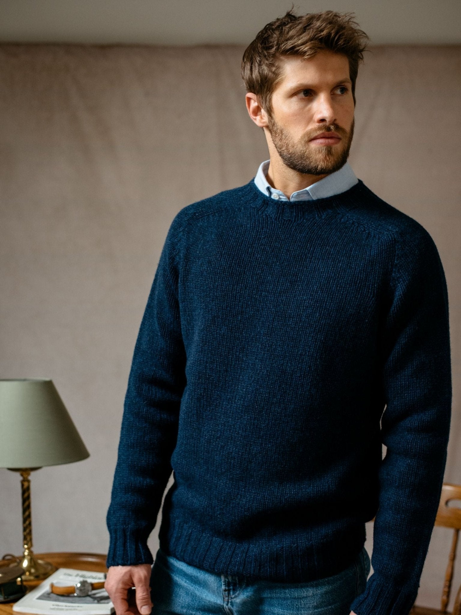 A man with short brown hair and a beard wears a dark blue Campbells of Beauly Geelong Lambswool Chunky Crew Jumper over a light collared shirt. Indoors by a table with a green lamp, he looks off to the side.