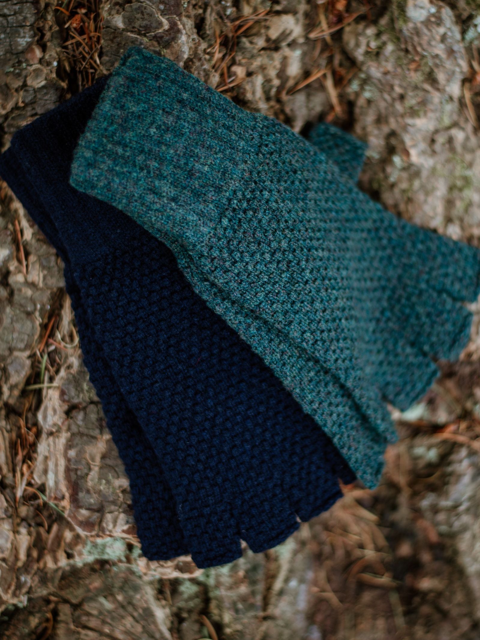 Two pairs of Campbells of Beauly Moss Stitch Half-Finger Gloves, one dark blue and one teal, rest on rough tree bark with pine needles—cozy winter essentials.