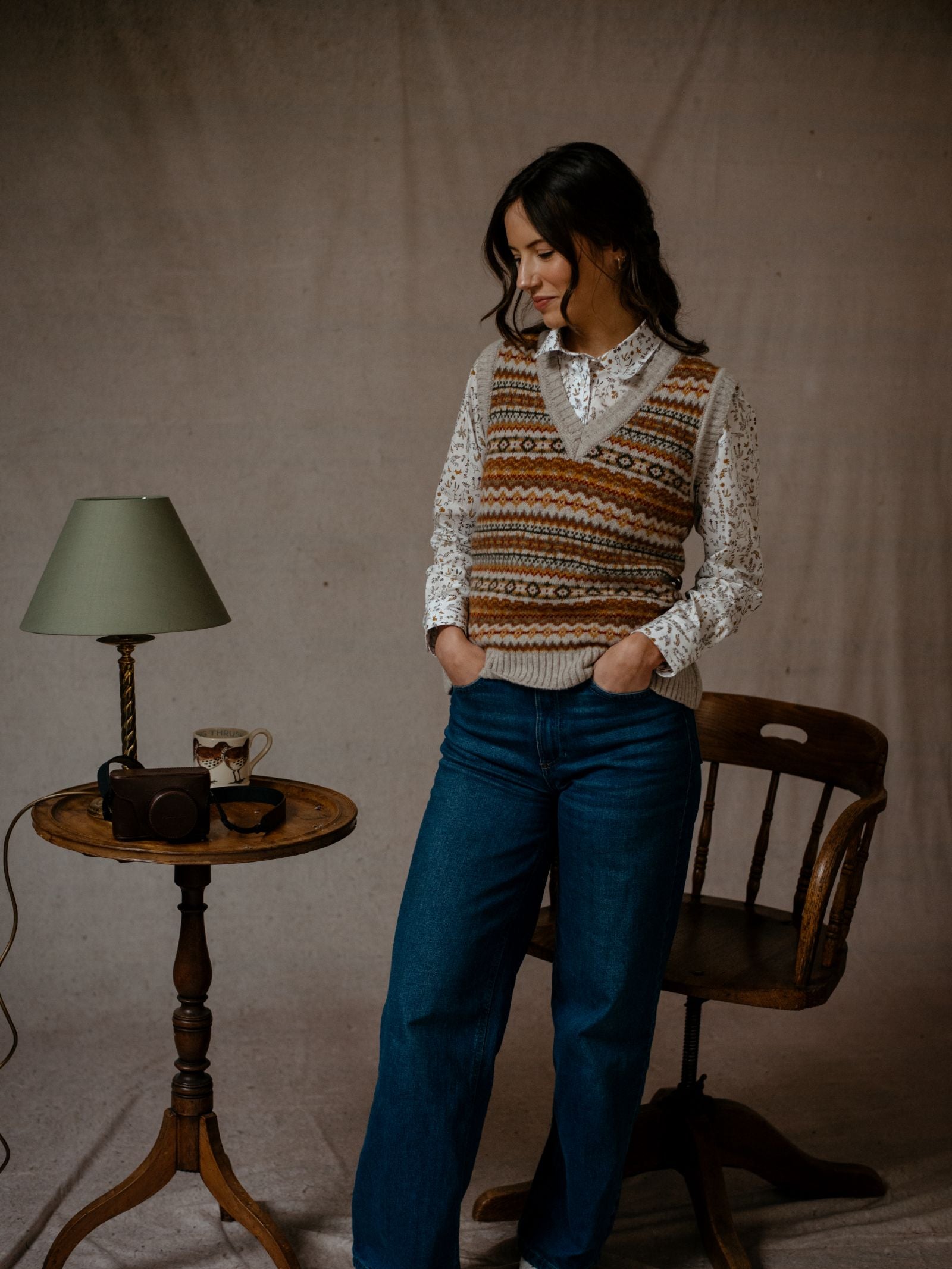 A woman in a Campbells of Beauly Natural Fairisle V-Neck Tank made from Shetland wool stands by a small round table with a lamp, mug, and camera. Hands in her jeans pockets, she smiles down slightly. A wooden chair sits nearby.