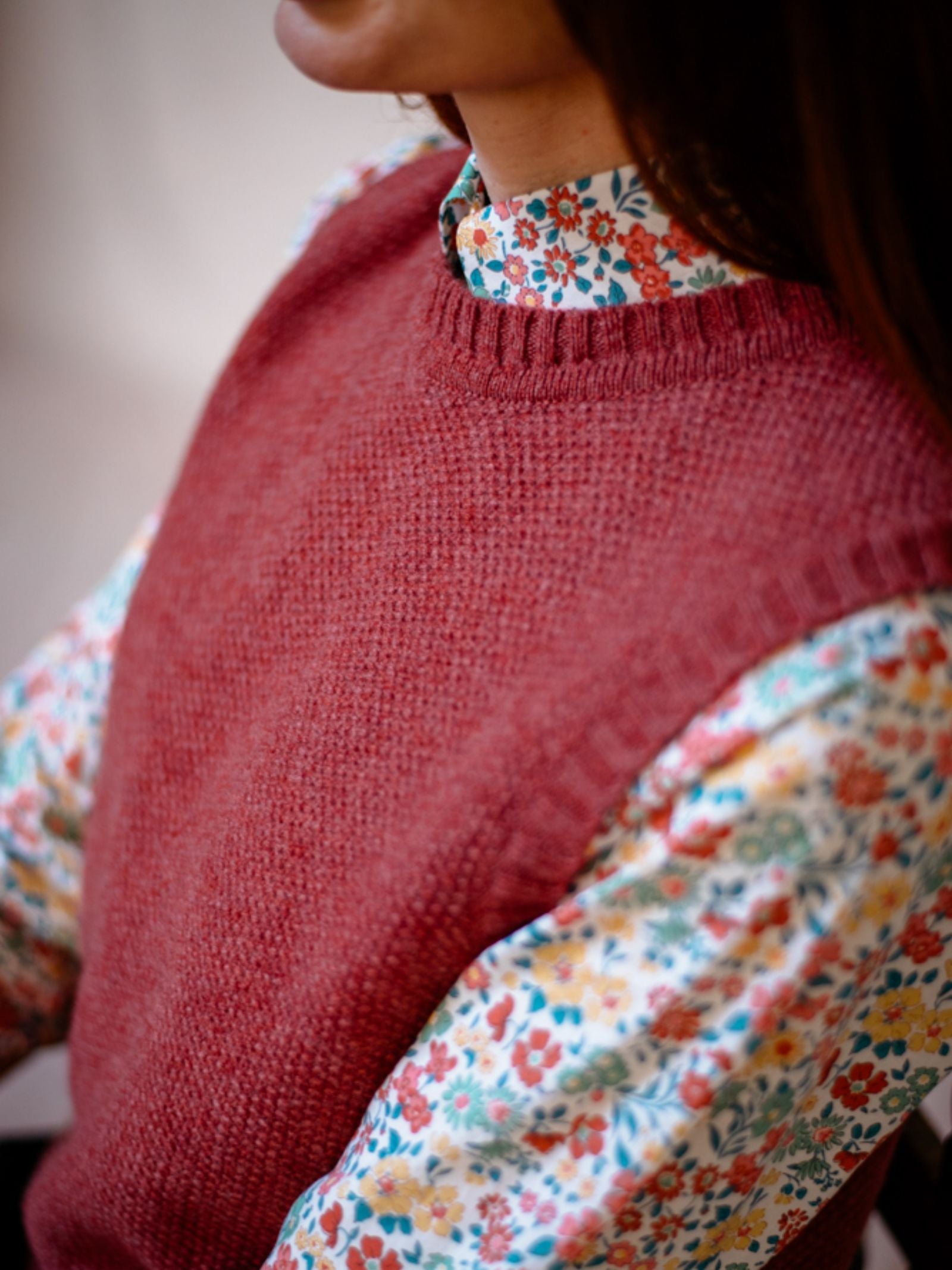 A person wears the Moss Stitch Crew Tank by Campbells of Beauly in red over a floral-patterned shirt with red, blue, and orange flowers. Made from Scottish yarn, this versatile top is shown in an upper body photo without the full face visible.