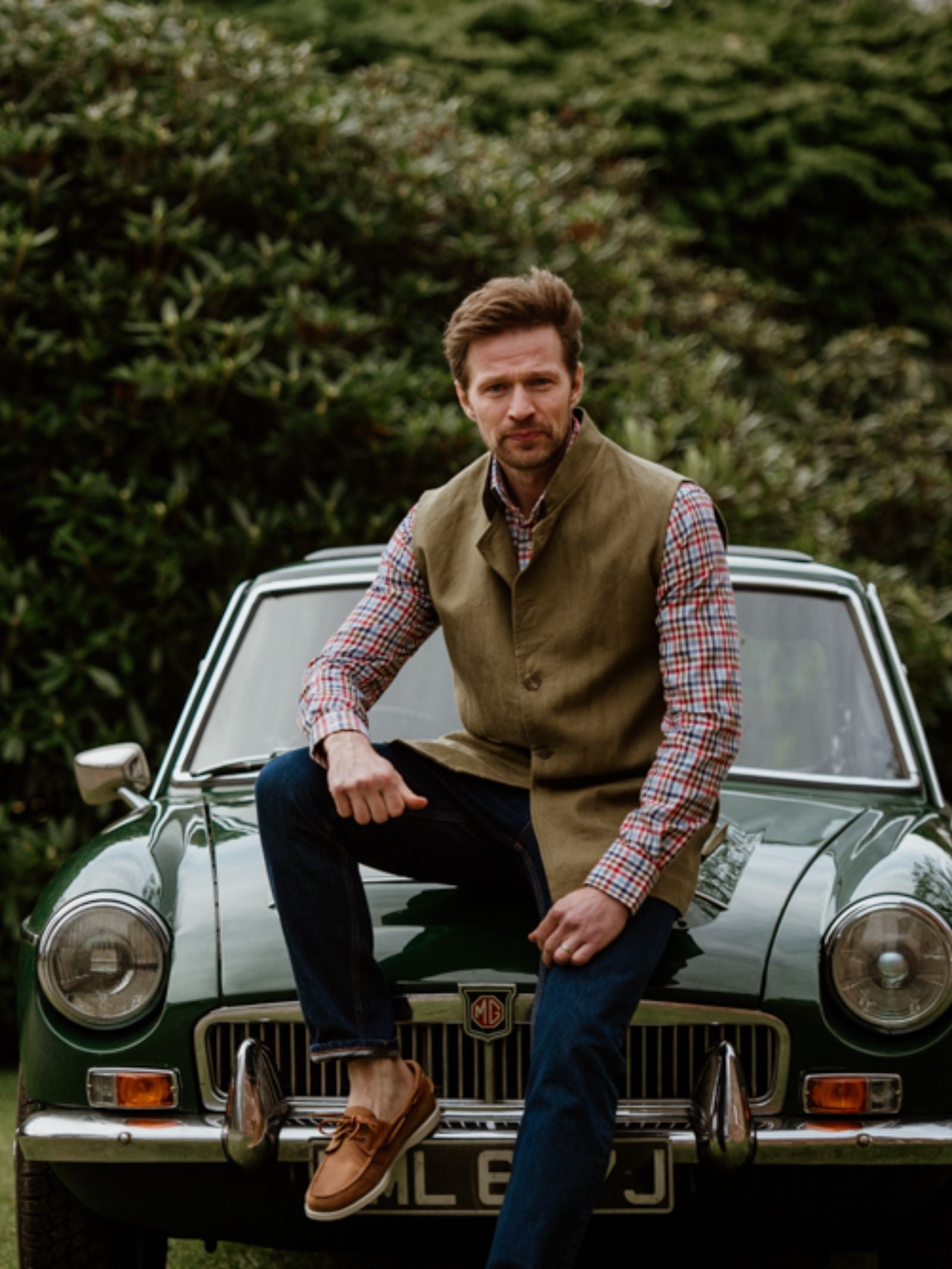 A man in a checked shirt, Campbells of Beauly Linen Nehru Gilet, jeans, and brown shoes sits on the hood of a vintage green MG sports car with greenery behind—a perfect summer layer.