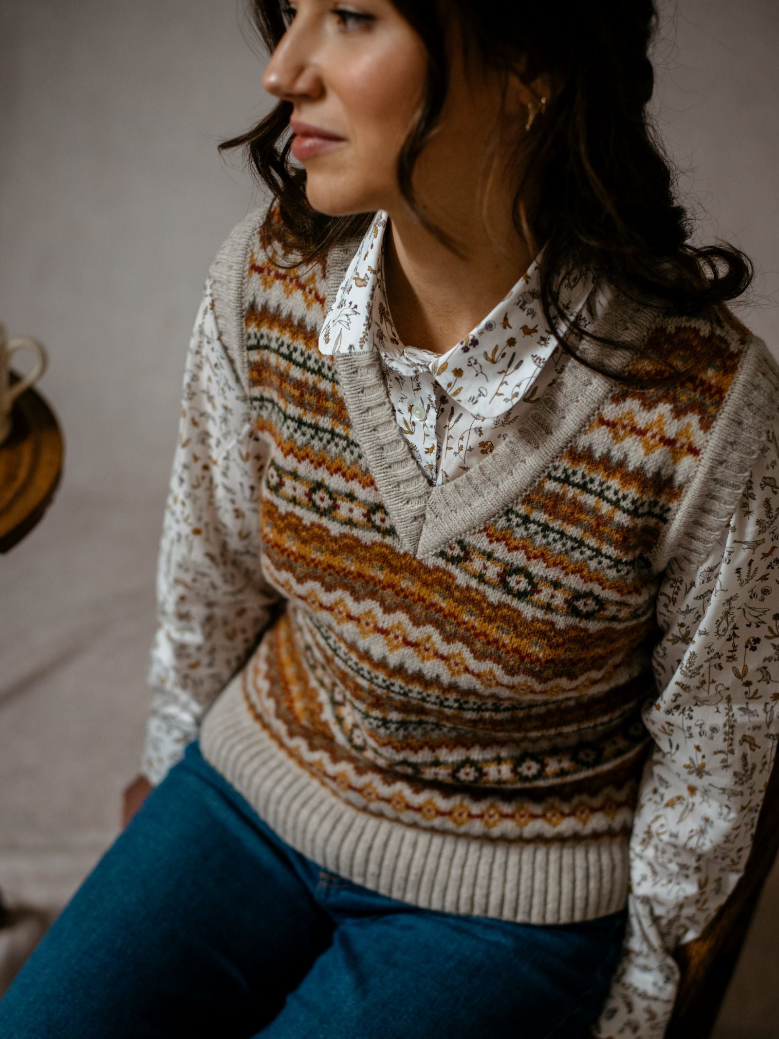 A woman with wavy brown hair, wearing Campbells of Beaulys Natural Fairisle V-Neck Tank over a white floral blouse and blue jeans, sits turned slightly to the side with a calm, relaxed expression.