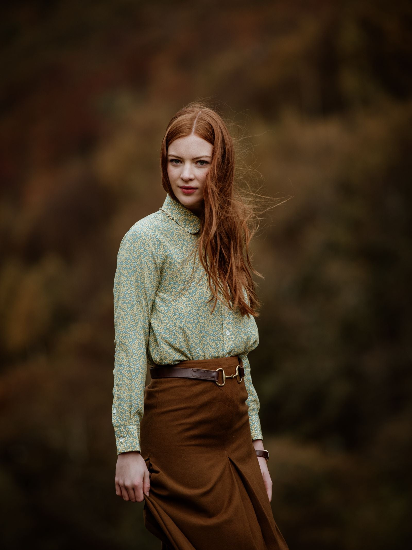A woman with long red hair wears a Campbells of Beauly Liberty Print Silk Shirt and a brown skirt while standing outdoors in front of blurred autumn trees.