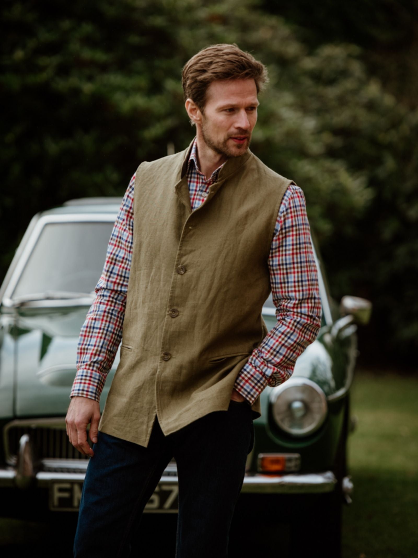 A man stands outdoors before a vintage green car, wearing Campbells of Beauly Linen Nehru Gilet over a red and white plaid shirt—a stylish summer layer with blurred greenery in the background.