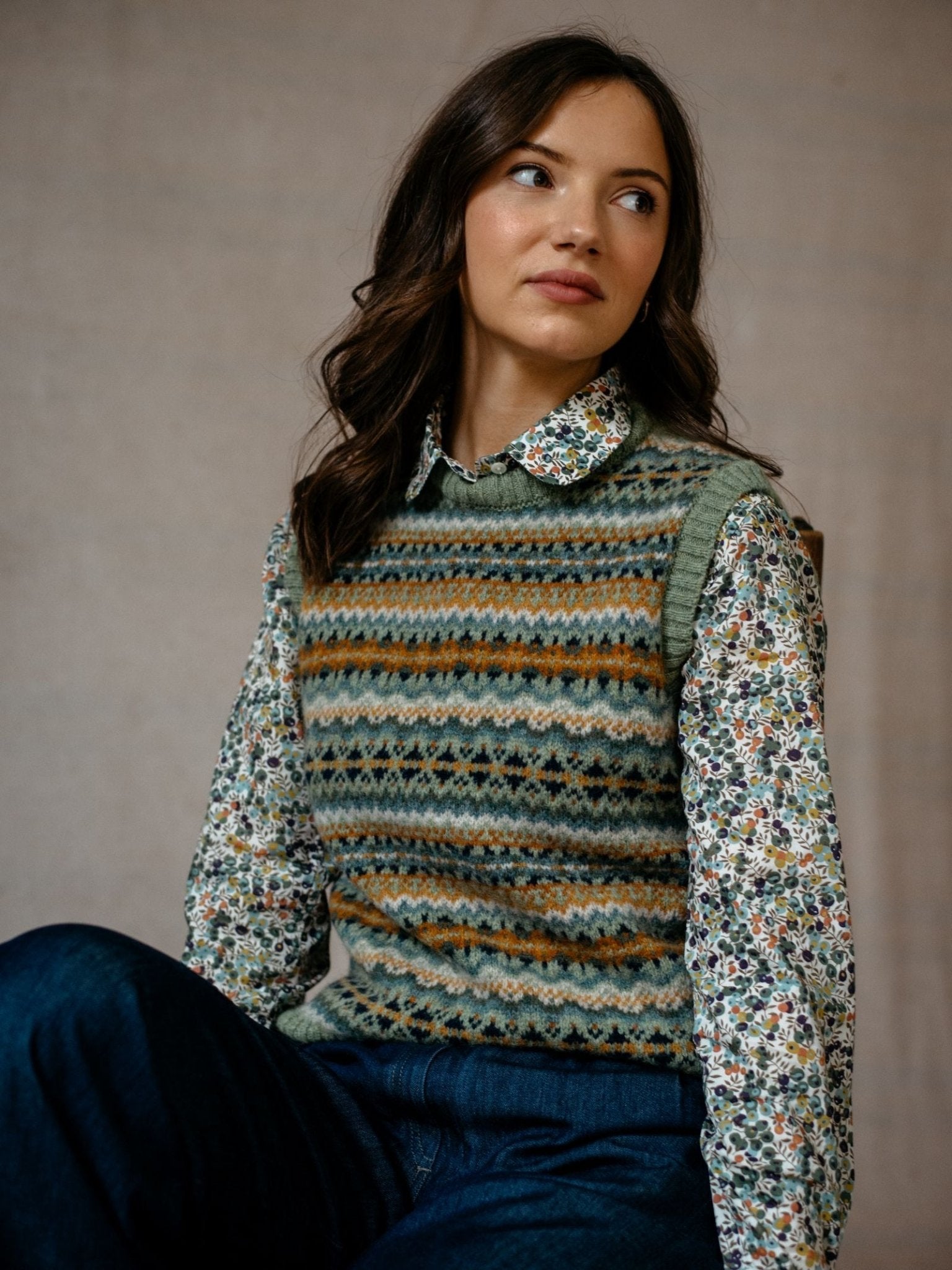 A woman with wavy brown hair sits against a neutral background, wearing Campbell's of Beauly Fairisle Crew Tank, knitted in Scotland from soft Shetland wool, over a floral collared shirt and dark jeans, looking slightly to her left.