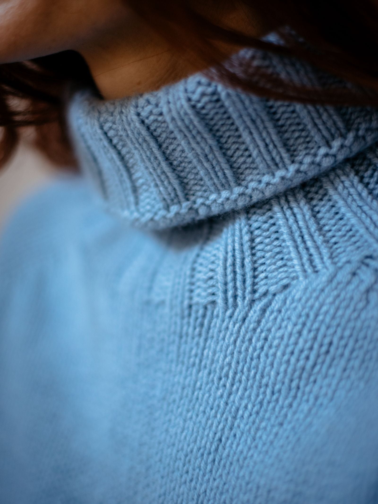 Close-up of a person wearing the Chunky Roll Neck Sweater by Campbells of Beauly in breathable, light blue ribbed knit. Brown hair frames the neck, highlighting the supersoft wool and cozy texture.