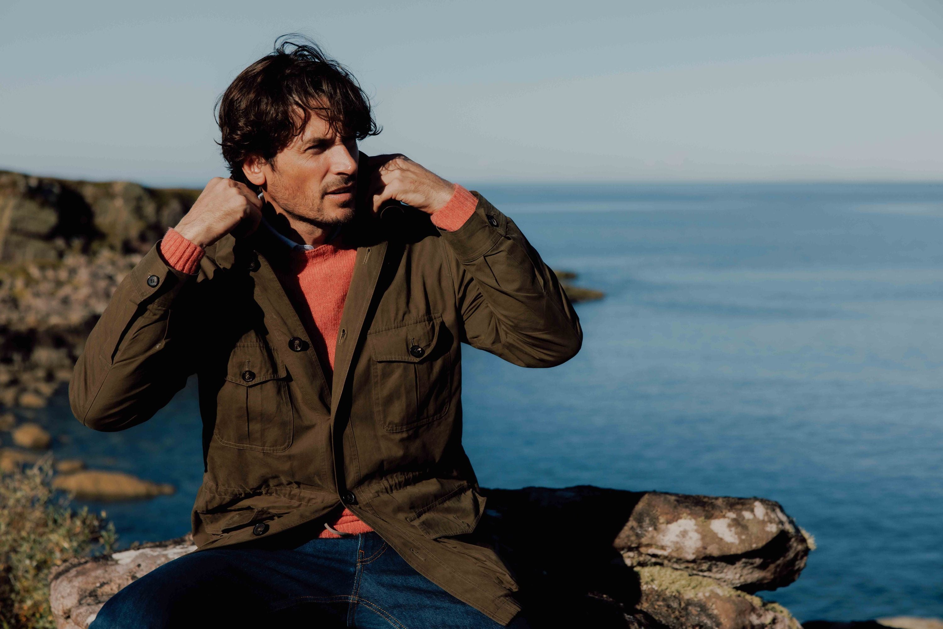 A man sits on a rock near the water, adjusting the collar of his olive green jacket. He is wearing a red sweater and blue jeans, with a calm sea and rocky shoreline in the background under a clear sky.
