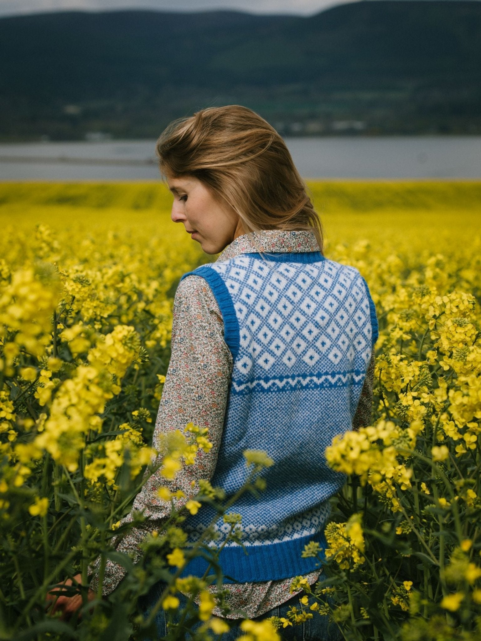 A woman stands in a field of yellow flowers, wearing the Campbells of Beauly Nordic V Tank over a floral shirt. Hills and a lake appear in the background beneath a cloudy sky.