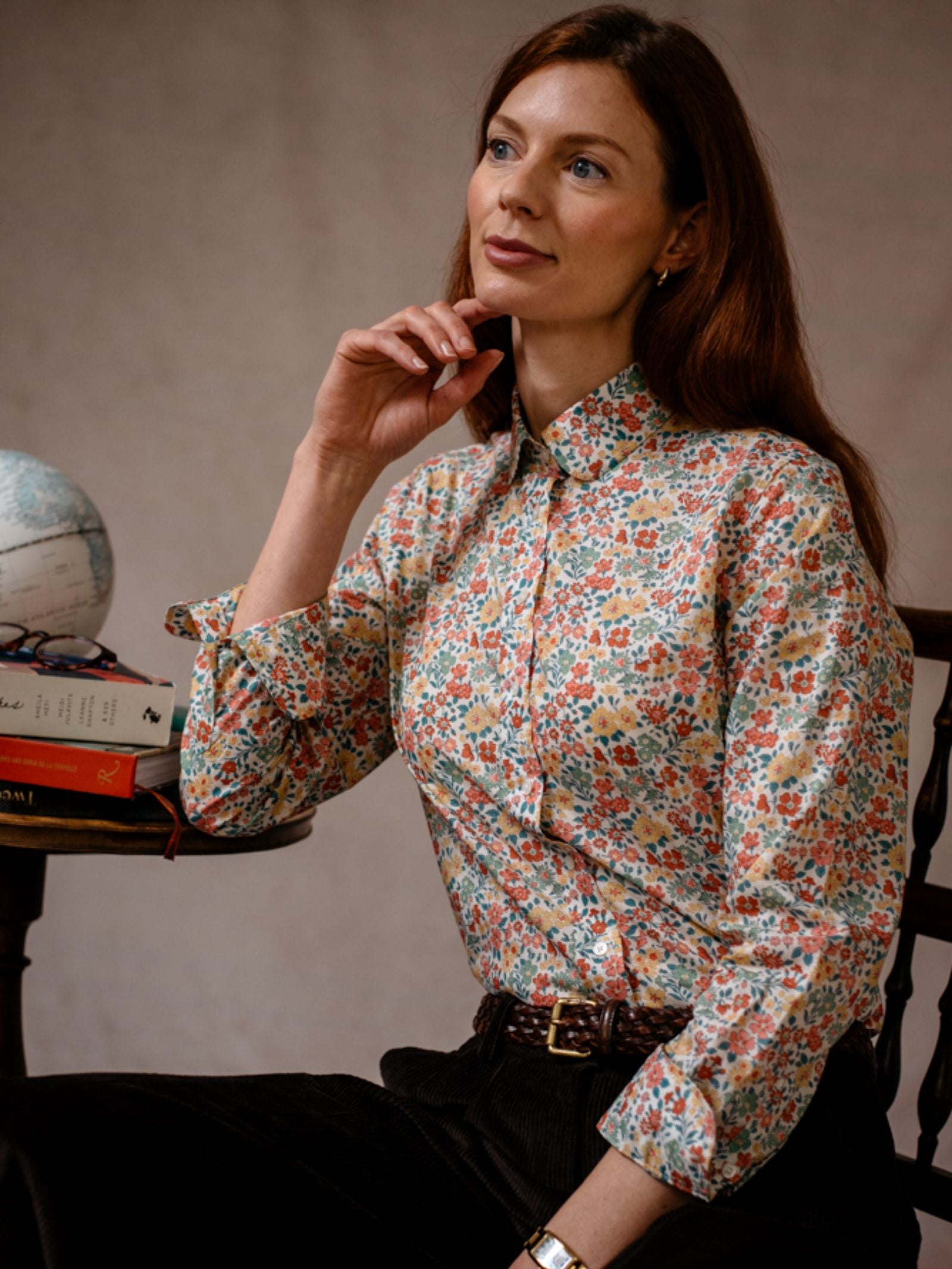 A woman with long brown hair wears a Campbells of Beauly Liberty Print Cotton Shirt and dark pants, sitting by a table with a globe and books, gazing thoughtfully into the distance.