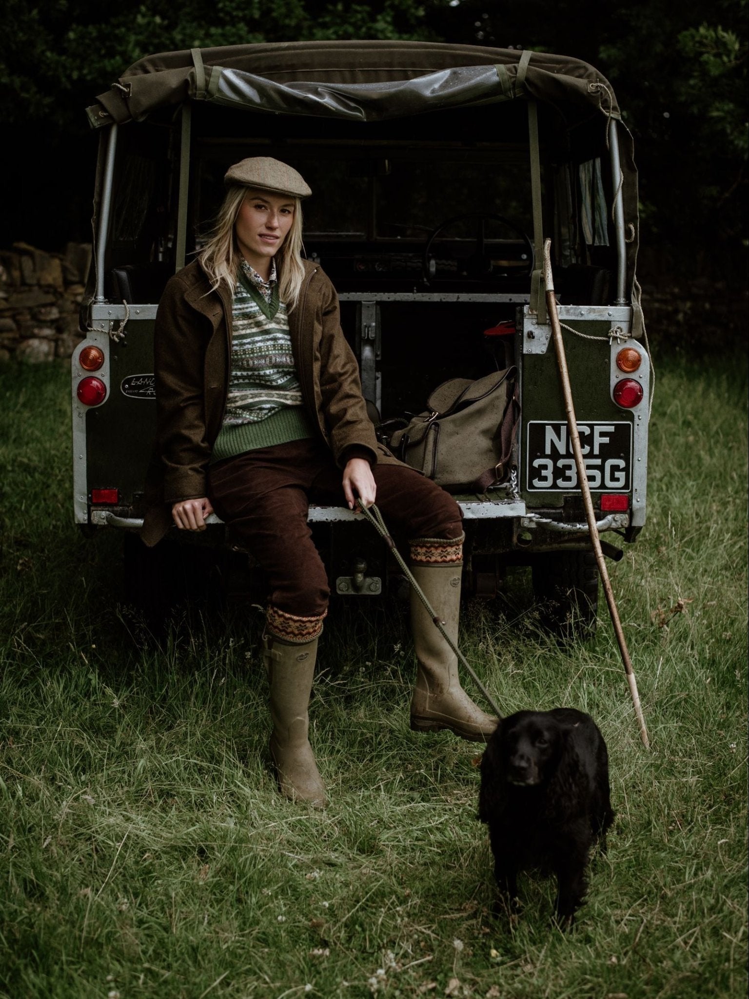 A woman in outdoor clothing and a Campbell's of Beauly Cashmere V-Neck Fairisle Slipover sits on a vintage Land Rover’s open tailgate in a grassy field, holding a leash with her black dog at her feet. Outdoor gear and bags are behind her.