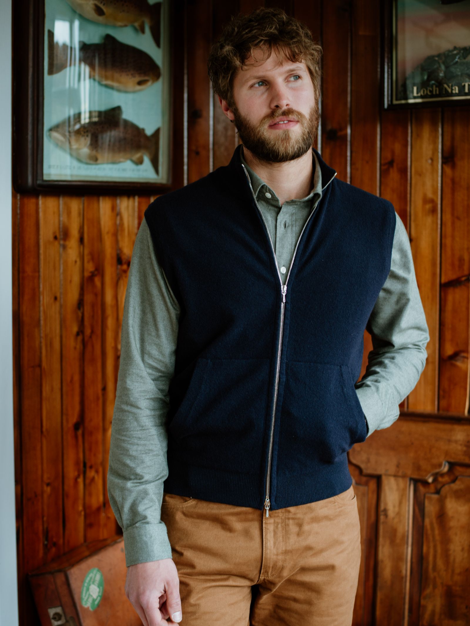 A bearded man wears a green button-up shirt, brown pants, and a navy Zip Gilet by Campbells of Beauly. He stands indoors against a wooden wall with framed fish illustrations behind him, exemplifying timeless style.