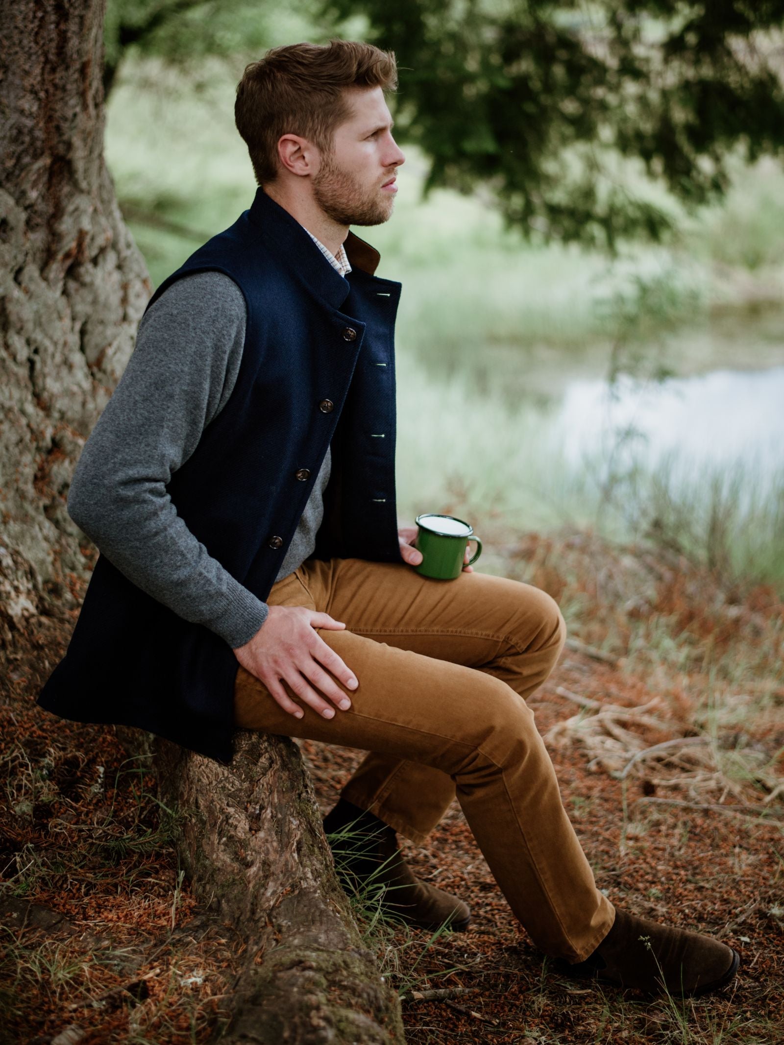 A man with light brown hair and a beard sits outdoors on a tree root, wearing tan pants, a gray sweater, and the Tweed Nehru Gilet by Campbells of Beauly. He holds a green mug; trees and a pond appear in the background.