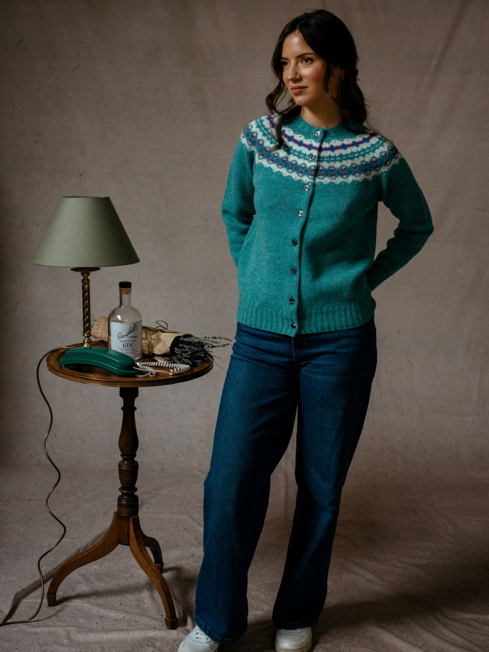 A woman wears the Campbells of Beauly Shetland Fairisle Cardigan with jeans, standing with hands behind her back next to a small round table holding a green lamp, bottle, glasses, books, and black purse against a neutral background.