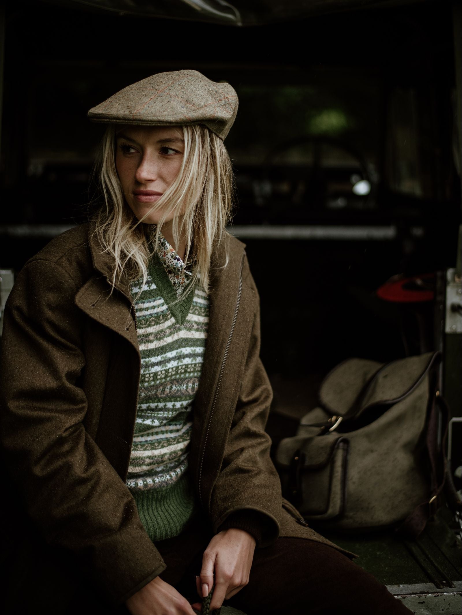 A woman in a Campbell's of Beauly Auld Stock Cashmere V-Neck Fairisle Slipover, tweed flat cap, and brown coat sits in the dimly lit back of a vehicle, looking to the side. A canvas bag rests beside her.