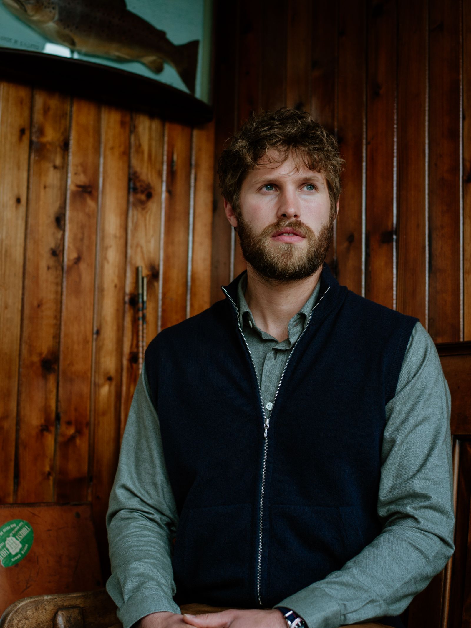 A man with curly hair and a beard sits indoors against a wooden wall, wearing a green shirt and the classic Zip Gilet from Campbells of Beauly. A mounted fish decorates the wall above him.