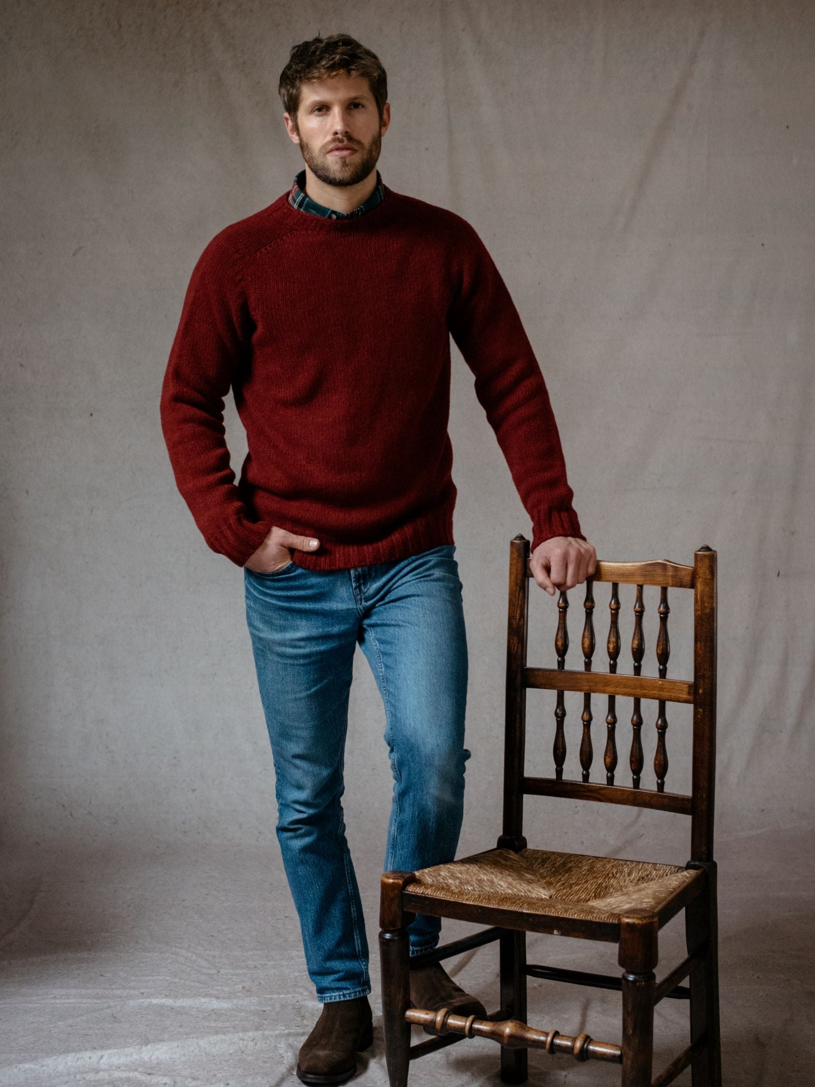 A man with short hair and a beard wears a red Campbells of Beauly Geelong Lambswool Chunky Crew Jumper, blue jeans, and brown shoes. He stands indoors with one hand on a wooden chair against a plain light background.