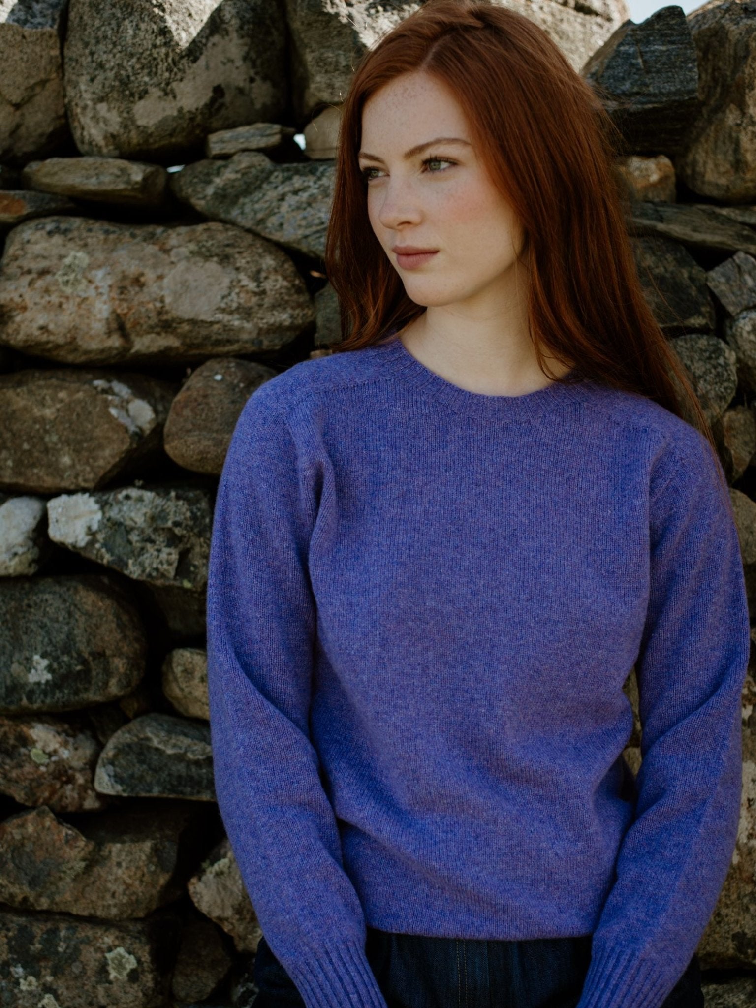 A young woman with long red hair, wearing a Campbells of Beauly Geelong Lambswool Crew Neck Jumper in purple, stands before a stone wall, gazing thoughtfully to the side.
