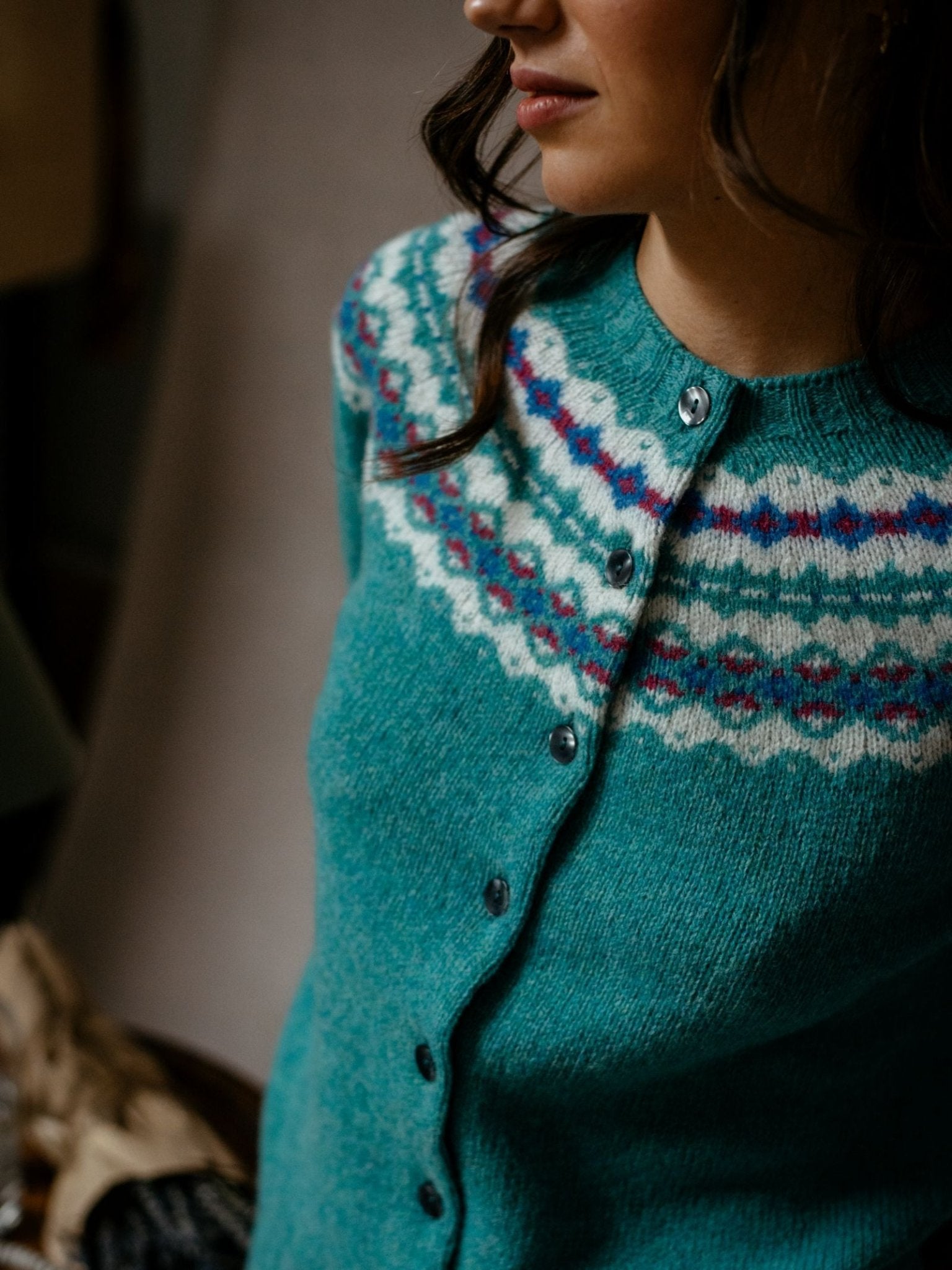 A woman wears the Campbells of Beauly Shetland Fairisle Cardigan in pure new wool, featuring a teal base and white, blue, and pink geometric yoke; only her upper body and part of her face are visible indoors.