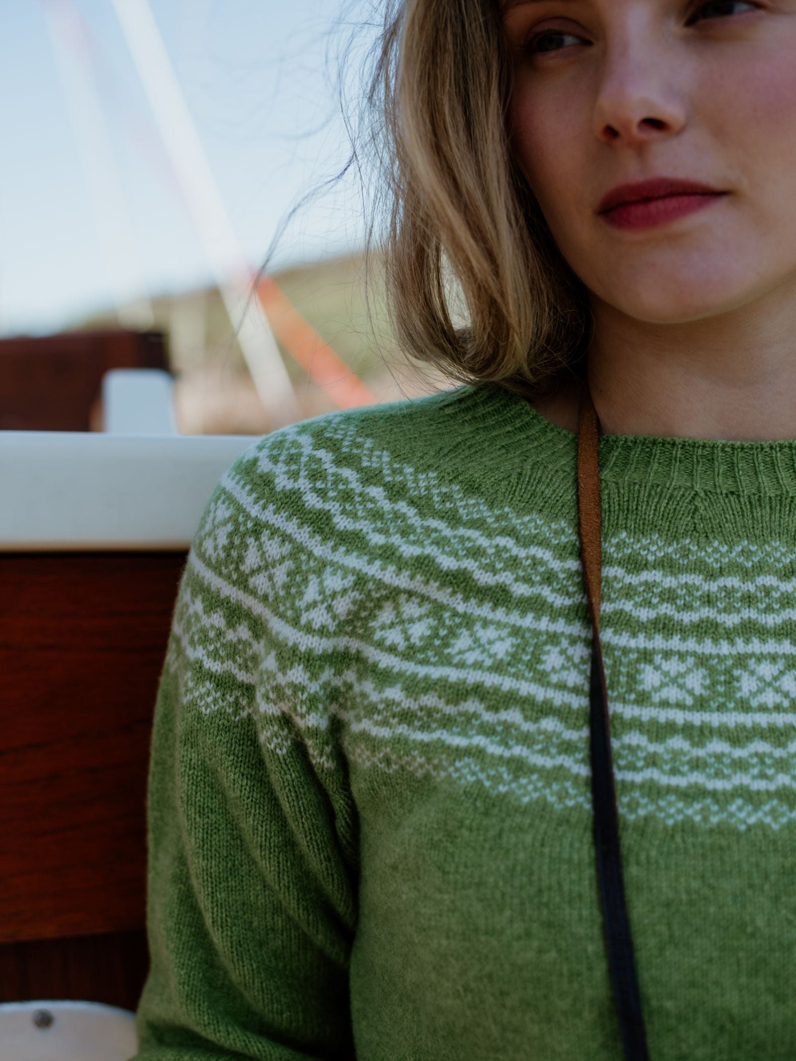 A woman with light brown hair wears Campbells of Beaulys Two-Colour Fairisle Yoke Jumper, featuring a white Nordic pattern, while sitting outdoors near a boat with wooden and white details—a nod to classic Scottish knitwear.