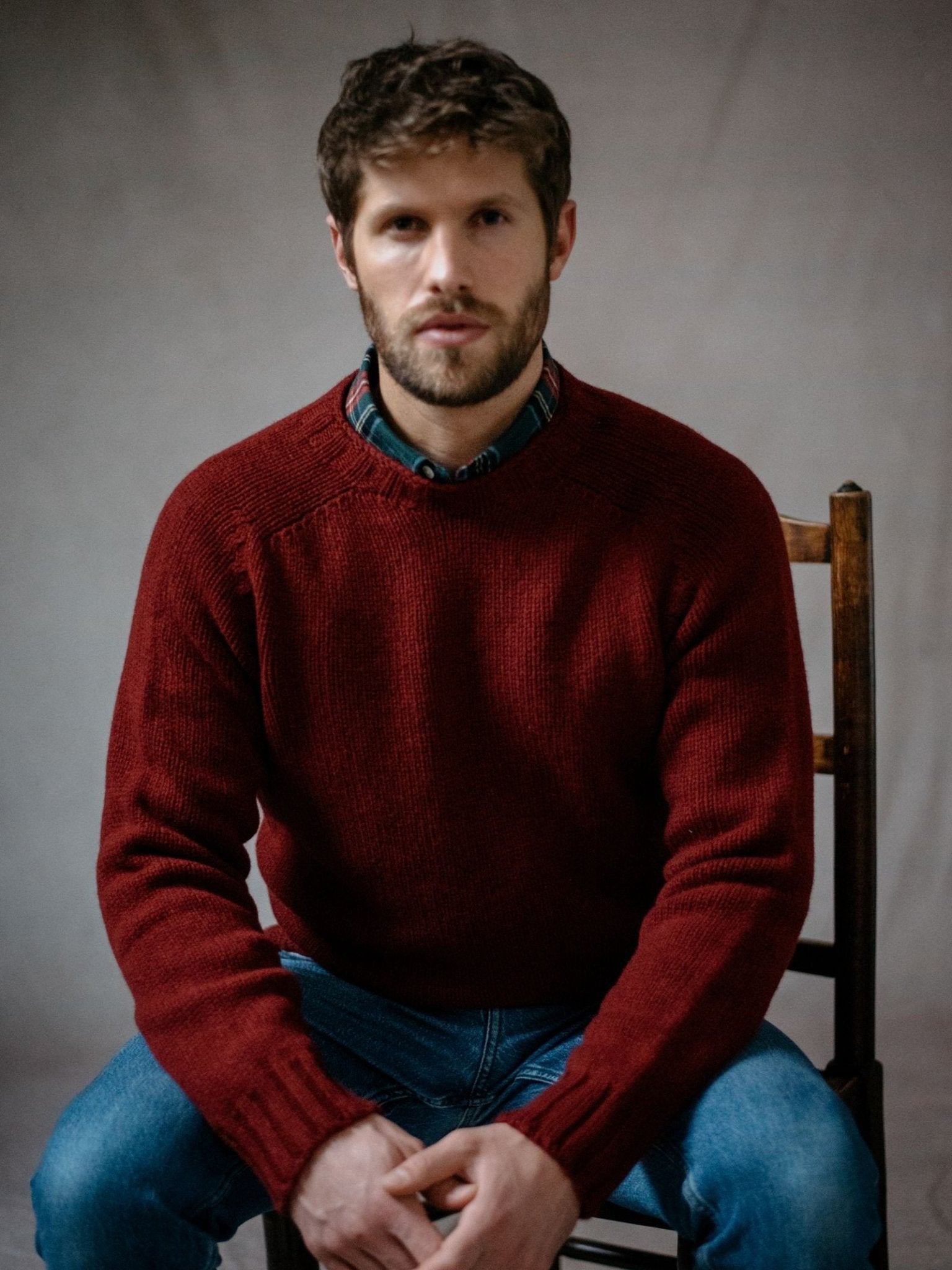 A man with short brown hair and a beard sits on a wooden chair, wearing a Campbells of Beauly Geelong Lambswool Chunky Crew Jumper in maroon over a plaid shirt and blue jeans, looking directly at the camera with a neutral expression.