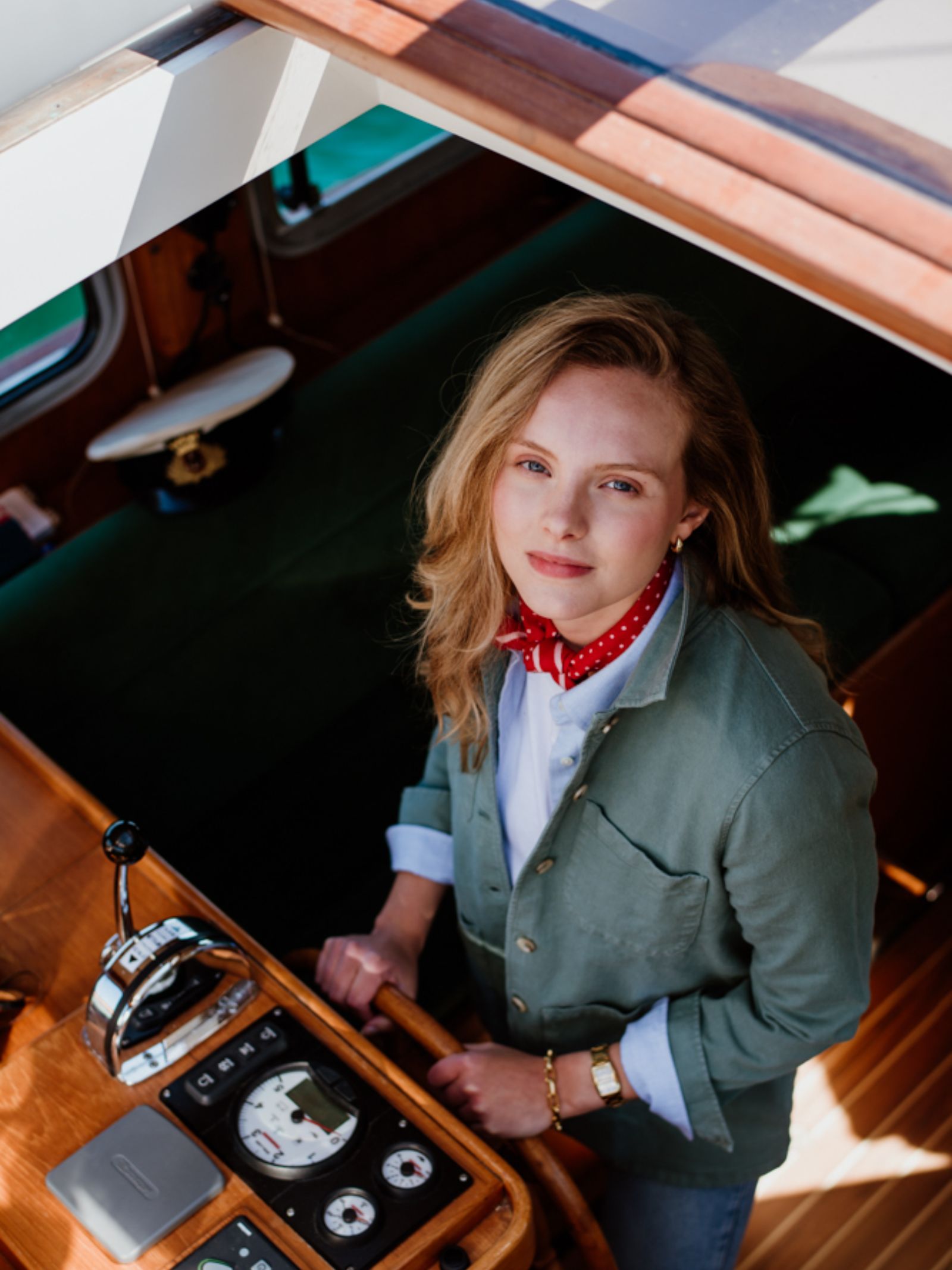 A blonde woman in a red polka-dot scarf and white shirt wears a relaxed fit green Campbells of Beauly Summer Chore Jacket at the helm of a wooden boat, sunlight illuminating the cozy cabin, boat controls, and captain’s hat.
