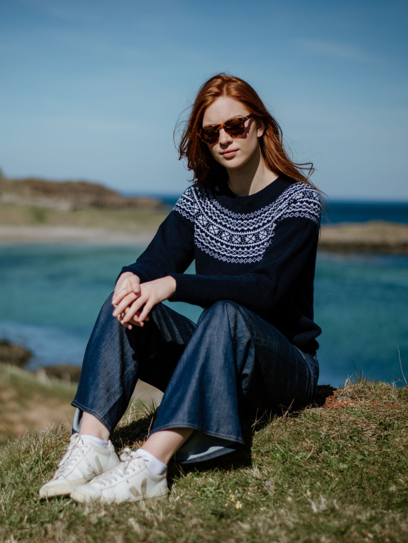 A woman with long red hair and sunglasses sits on grass by the sea, wearing a Campbells of Beauly Two-Colour Fairisle Yoke Jumper, wide-leg jeans, and white sneakers, with blue sky and water in the background.